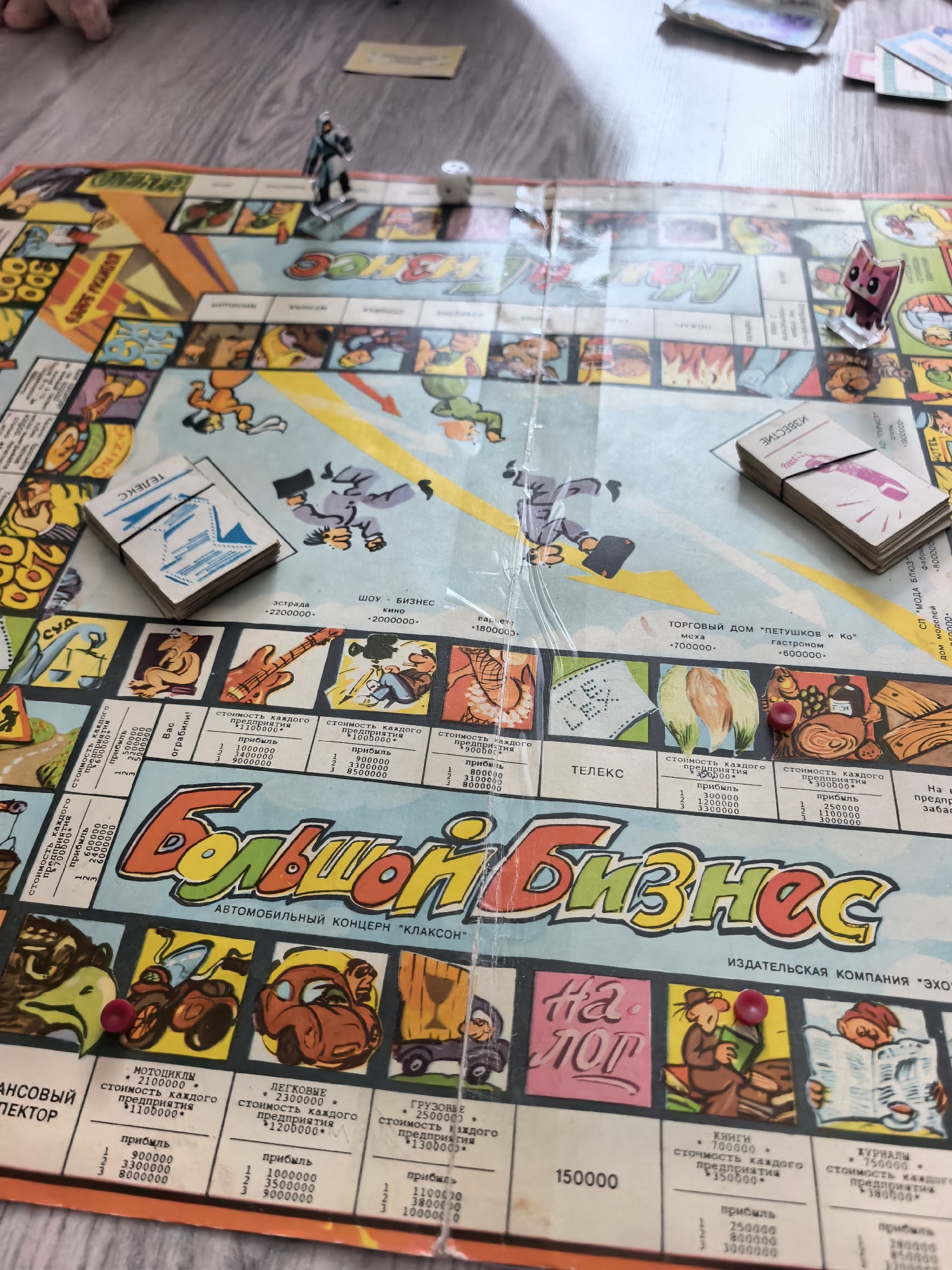 The image shows a close-up, overhead view of a board game, likely Monopoly, with a focus on the game board and some scattered game pieces. The board is colorful and features various property names and illustrations in Russian, including "Большой Бизнес" (Big Business) as the title. Several game tokens, including a small human figure, a pink cat-like figure, and a red die, are visible on the board. Some paper money or property cards are also present. The game is set on a wooden surface, suggesting it is being played on a table. The lighting is natural, coming from above and slightly to the right. The image is sharp and well-focused, providing clear detail of the game's elements. There is no discernible connection to the city of Sterlitamak, Russia.