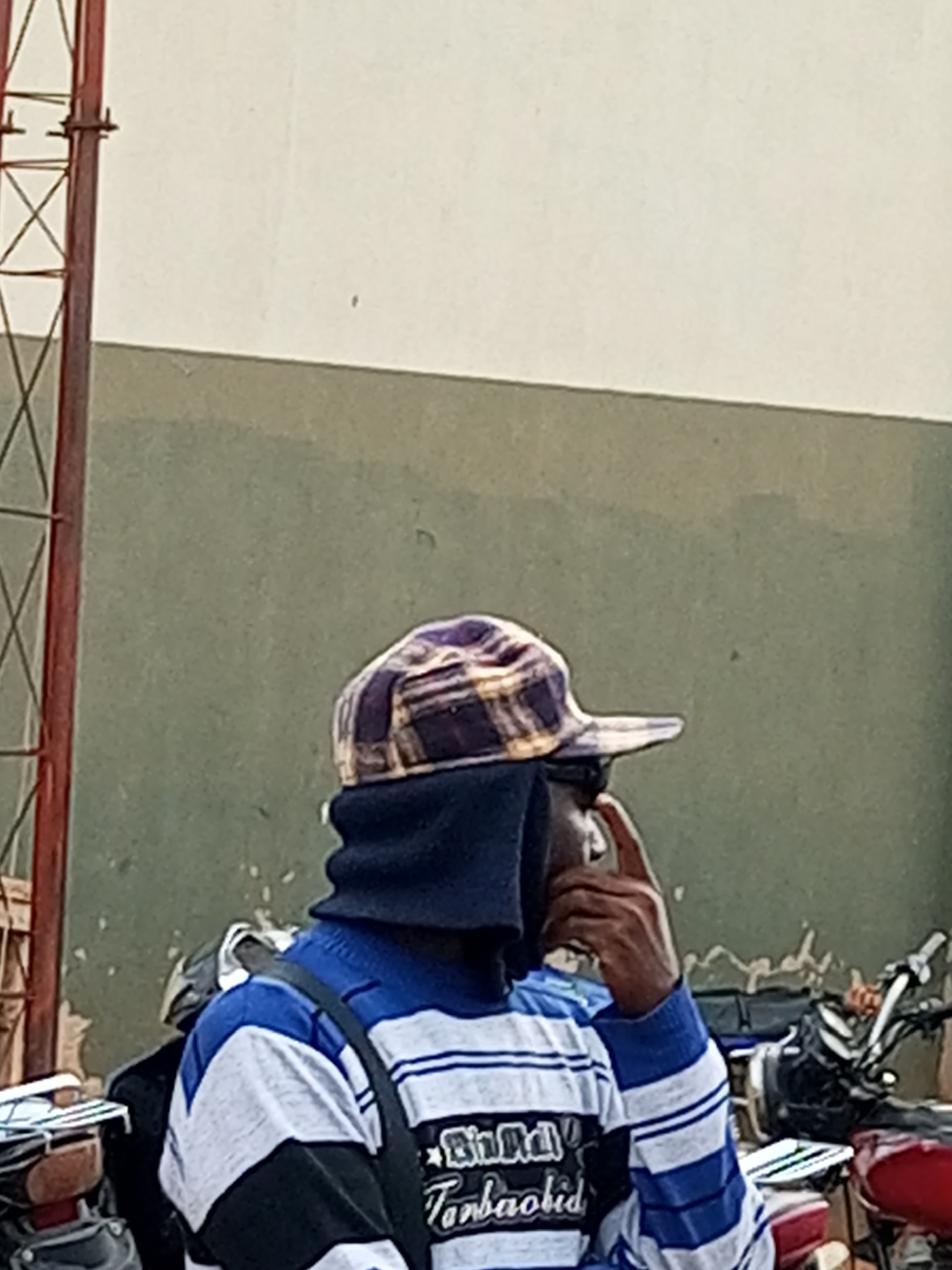 The image shows a person wearing a plaid cap and a blue and white striped long-sleeved shirt. The person is also wearing a dark neck gaiter or scarf that covers their lower face. They are seen from the chest up, with their right hand raised to their face, appearing to be touching their nose or chin. In the background, there are motorcycles and a textured wall. The setting appears to be outdoors, possibly in a marketplace or a street in Gombe, Nigeria. A red metal structure, possibly part of a building or scaffolding, is visible on the left side of the image.