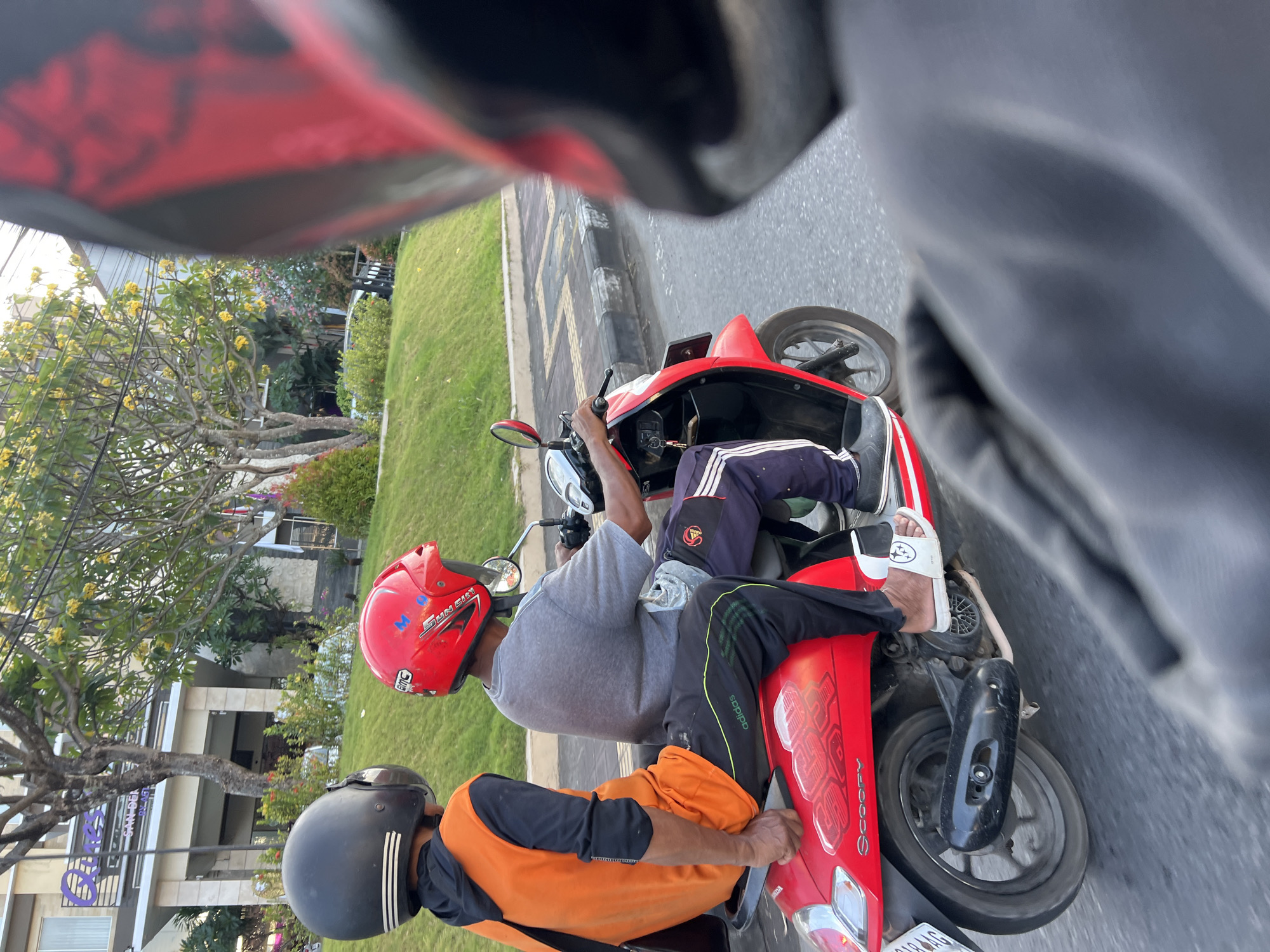 Two individuals are riding a red Honda Scoopy motorcycle on a paved road in Denpasar, Indonesia. The person in front is driving the motorcycle and is wearing a gray t-shirt, dark pants with white stripes, and white sandals. They have a red helmet. The passenger behind is wearing an orange vest over a dark long-sleeved shirt and dark pants, and a black helmet. They are holding onto the driver. The motorcycle has a license plate with the number "AG" visible. To the left of the road is a grassy embankment and a building with a sign that reads "Quinci Sanur". Trees with yellow flowers are visible in the background. The perspective suggests the photo was taken from another vehicle, possibly a motorcycle, following behind. The lighting indicates it is daytime.