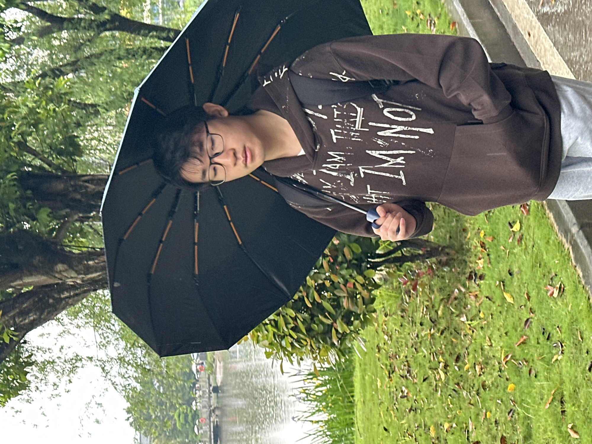 A young Asian male with glasses is holding a black umbrella while standing outdoors in Jiangjin, China. He is wearing a brown hoodie with white writing and grey sweatpants. The background includes trees, grass, fallen leaves, a body of water, and a grey concrete curb.