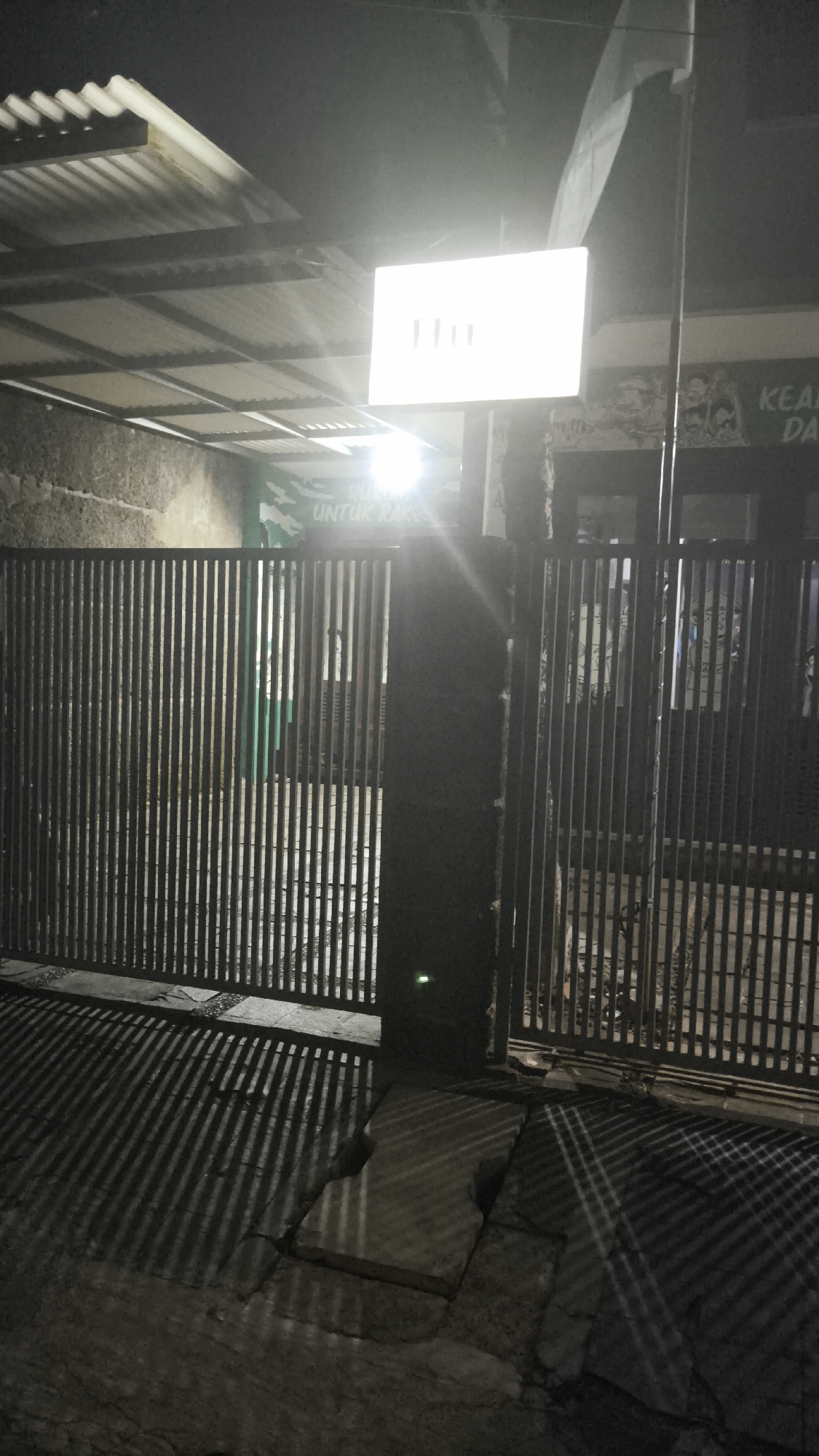 The image depicts an outdoor scene at night, possibly in South Jakarta, Indonesia. A corrugated metal roof extends over a paved area. In the center, a brightly lit rectangular sign with vertical lines is mounted on a pole. To the left and right, tall, dark metal gates are partially open. Behind the left gate, a green banner with white text that reads "UNTUK RAKYAT" is visible. The ground in front of the gates is paved, with cracks and uneven surfaces, and exhibits strong striped shadows cast by the gates. The overall impression is one of a dimly lit street or entryway, possibly a business or residential complex.