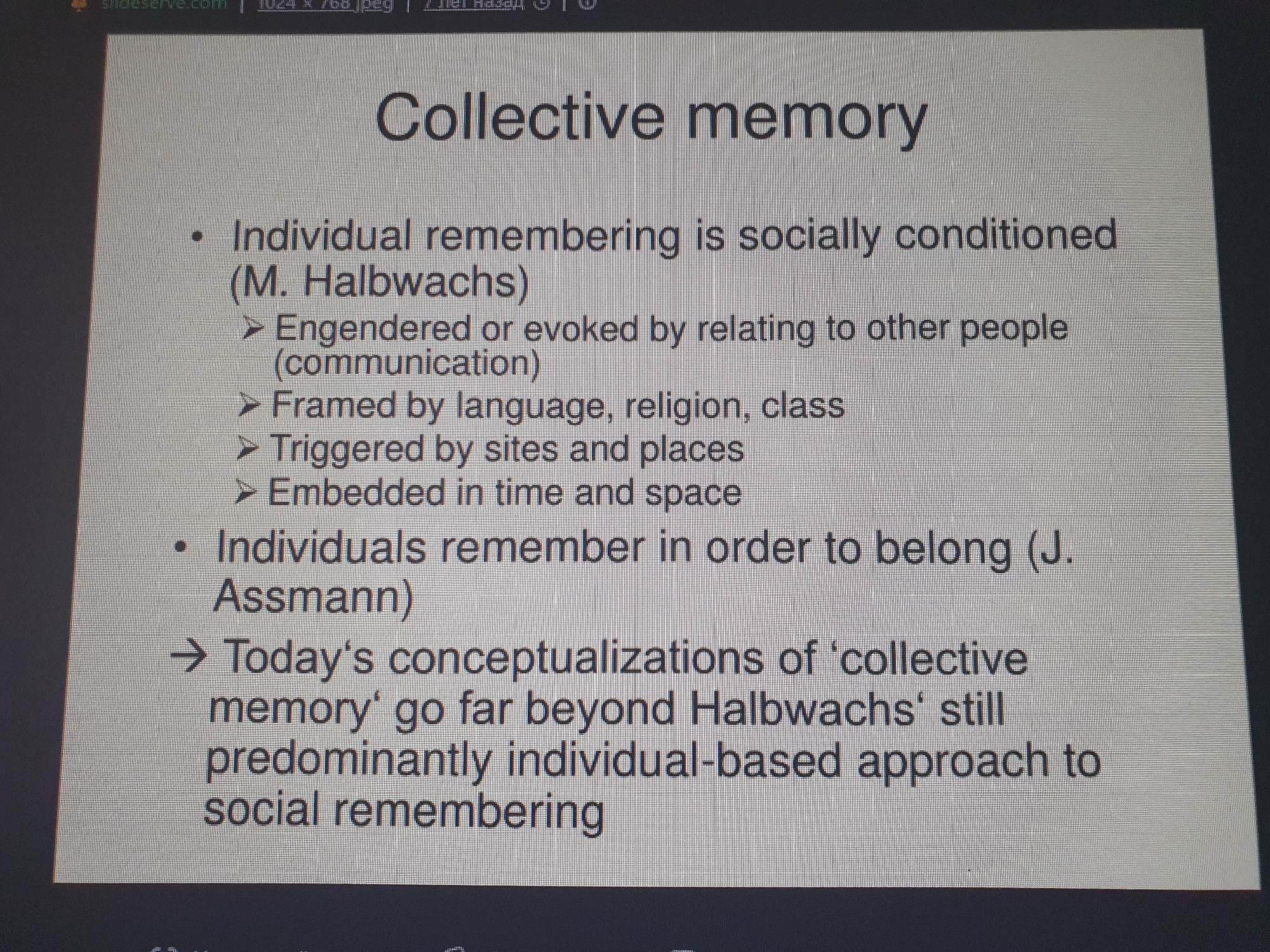 The image shows a projected presentation slide with text about "Collective memory". The text is centered on a light grey background. The slide title "Collective memory" is at the top. Below the title, there are bullet points and sub-points. The first bullet point states "Individual remembering is socially conditioned (M. Halbwachs)" and has three sub-points indicated by arrows: "Engendered or evoked by relating to other people (communication)", "Framed by language, religion, class", and "Triggered by sites and places". There is a second bullet point that reads "Individuals remember in order to belong (J. Assmann)". Finally, there is a final point indicated by a larger arrow: "Today's conceptualizations of 'collective memory' go far beyond Halbwachs' still predominantly individual-based approach to social remembering". The top left corner of the slide has a partial view of a website address: "slideserve.com". There are no people or any discernible environmental elements from the city of Tashkent, Uzbekistan, present in the image.