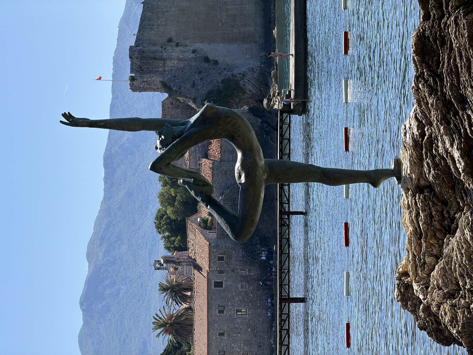 The image depicts a bronze statue of a gymnast in a contorted pose in the city of Budva, Montenegro. The statue is located on a rocky outcrop next to a body of water with swimming lane markers. In the background, a stone building with a red tiled roof, palm trees, and mountains are visible. On a pier adjacent to the statue, two people are standing, one appearing to dive into the water. In the far background, there is a fortress wall, with a red flag on top. A vapor trail is visible in the sky.
