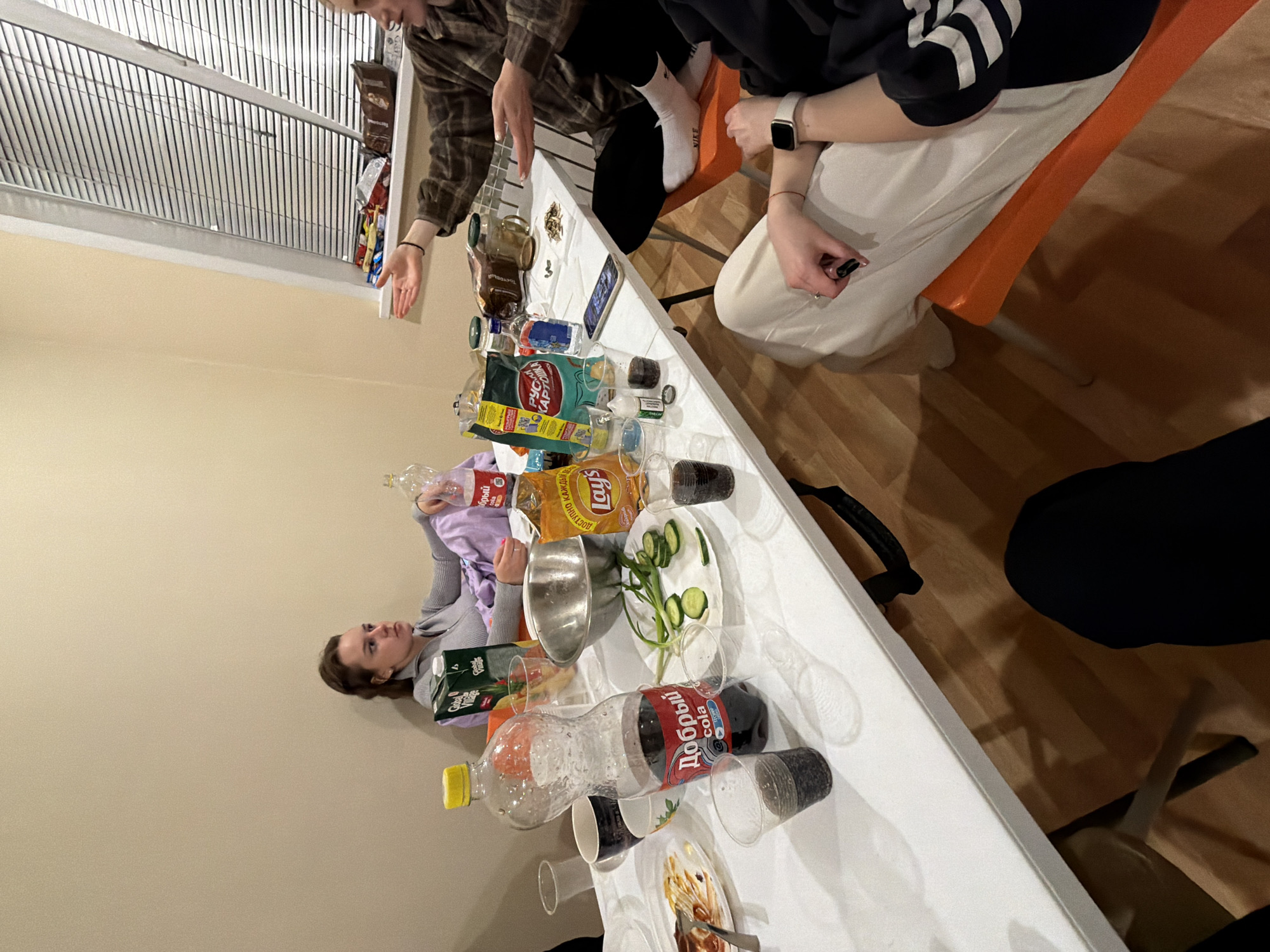 A medium shot captures a group of individuals gathered around a table, likely in a domestic setting. The table is laden with various items, including food, drinks, and disposable cups. The scene suggests a social gathering or party.
People: Four people are partially visible. One woman is seen in profile, lying on a lilac-colored hoodie.
Objects: Items visible on the table include bottles of soda, empty plastic bottles, a carton of Club Village tomato juice, a bag of Lays potato chips, a metal bowl, a plate of sliced cucumbers, and disposable cups.
The city of Perm in Russia is not evident from the image.