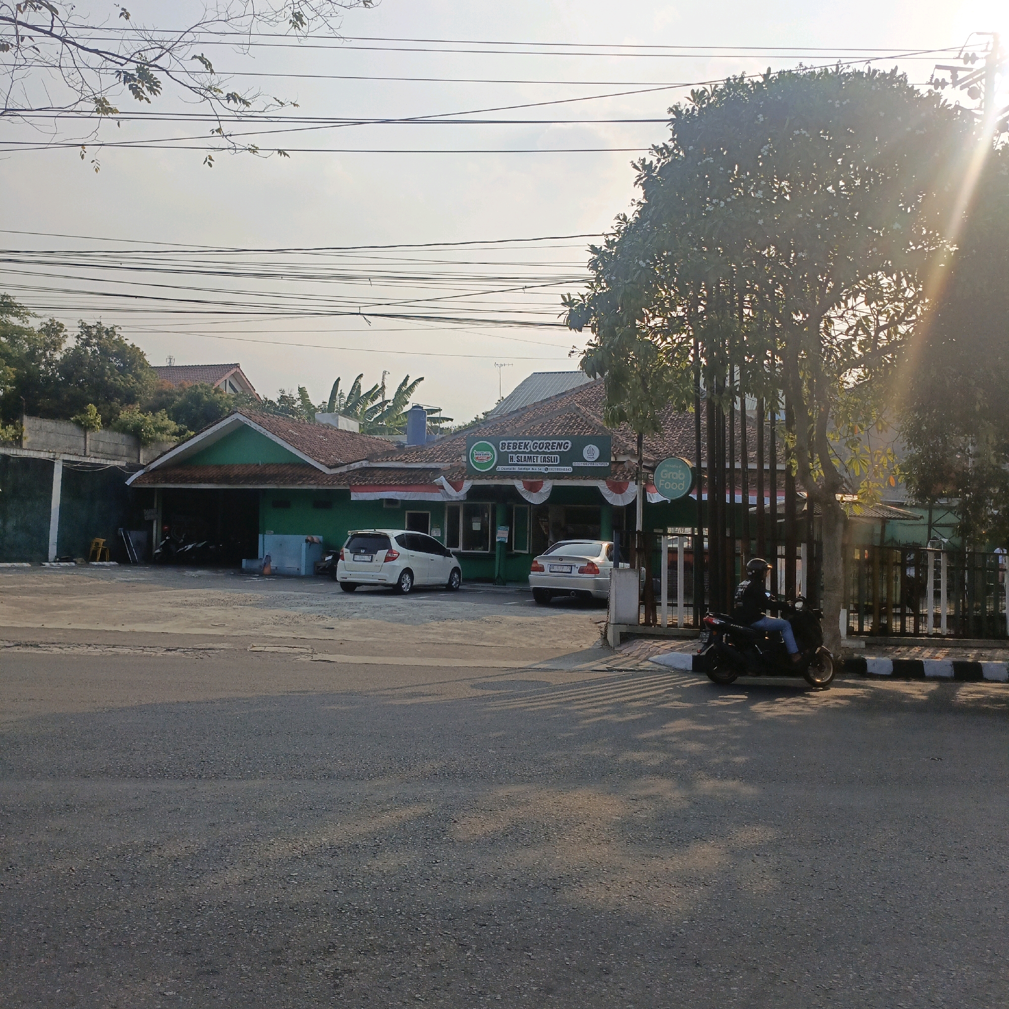 The image captures a street view in Salatiga, Indonesia, on a bright, sunny day. A green building with a red tile roof, identified by a sign as "BEBEK GORENG H. SLAMET (ASLI)", is prominently featured. The establishment appears to be a restaurant, with several cars parked in front, including a white Honda Jazz and a silver sedan. A GrabFood sign is visible on a fence to the right of the building. A person is riding a black motorcycle along the road, wearing a helmet and blue jeans. The road surface is asphalt, and there are shadows cast by overhead wires and trees. The environment includes trees, power lines, and residential buildings in the background. The sun is shining brightly, creating lens flare in the upper right portion of the image.