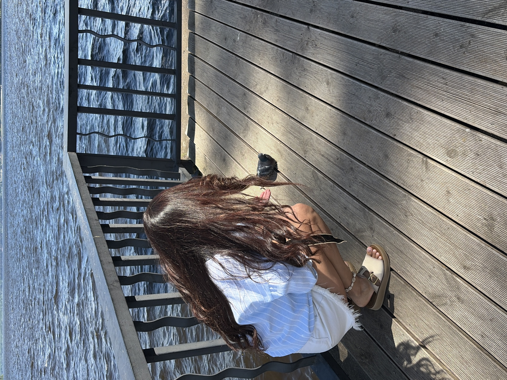 A woman with long, dark hair is sitting on a wooden deck next to a body of water. She is wearing a light blue and white striped shirt and white shorts. She has on tan platform sandals. The deck is made of wooden planks that run diagonally across the frame. The water is choppy and reflects the sunlight. There are metal stairs leading down to the water on the left side of the image. A pigeon is perched on the deck near the woman's feet. The sun is casting shadows on the deck, indicating it is daytime. The setting appears to be an outdoor area, possibly a park or waterfront promenade, in Chaikovsky, Russia. The overall mood seems relaxed and peaceful. There is no visible text in the image.