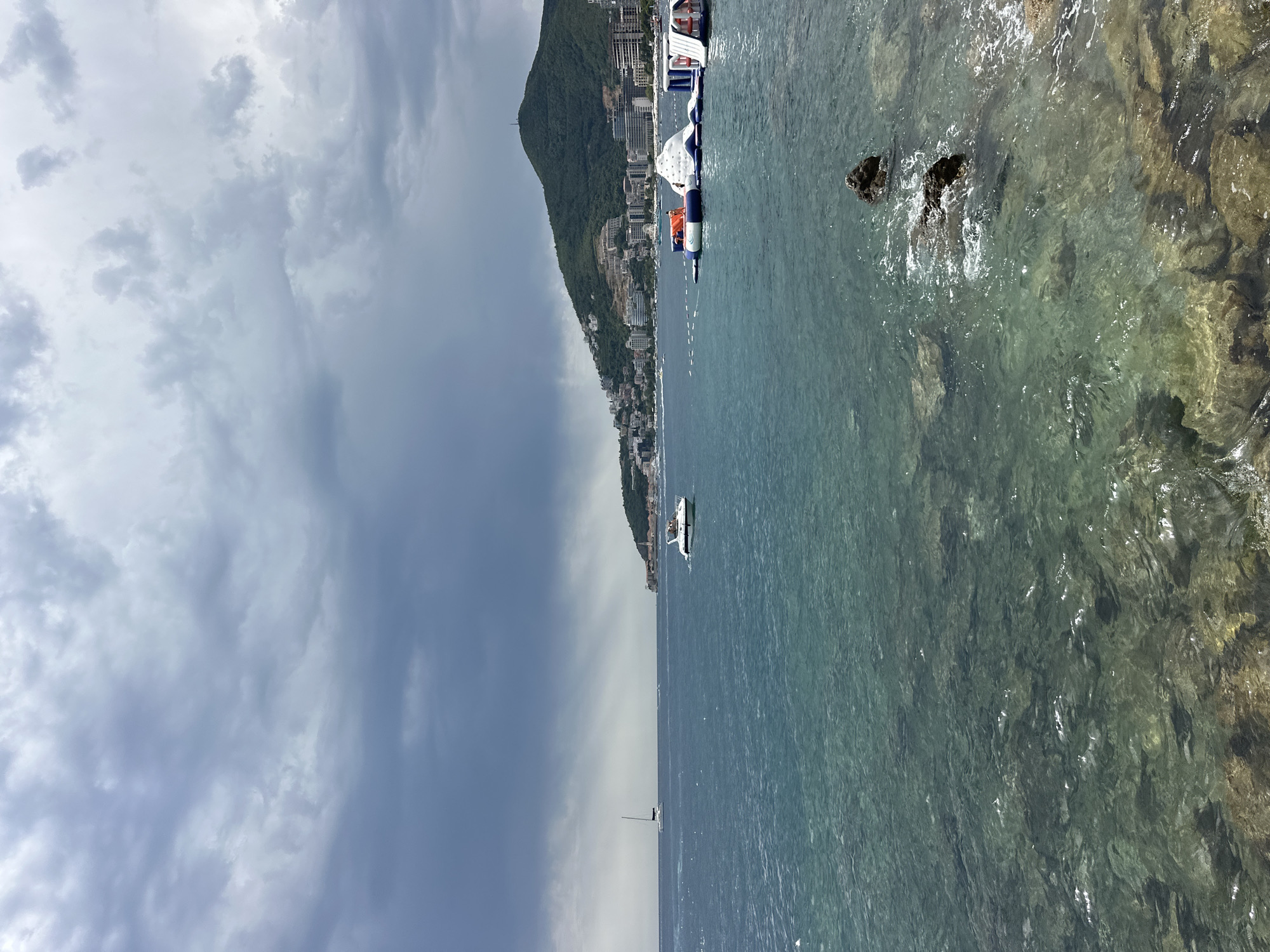 This is a horizontal, eye-level, medium shot of the Budva Riviera in Montenegro, taken from the water's edge. The scene is dominated by a choppy, turquoise sea under a dark, cloudy sky, indicating an impending storm. In the foreground, rocks are visible partially submerged in the shallow water. Further out, a small white boat with several occupants sails across the frame, and a white sailboat is seen in the distance. Along the coastline, a densely populated city, Budva, sprawls across a green, mountainous backdrop. A prominent inflatable water park, featuring various structures like slides and climbing walls, is situated in the water to the right of the frame. The water park has orange and white inflatable elements, with at least one person visible on it.
