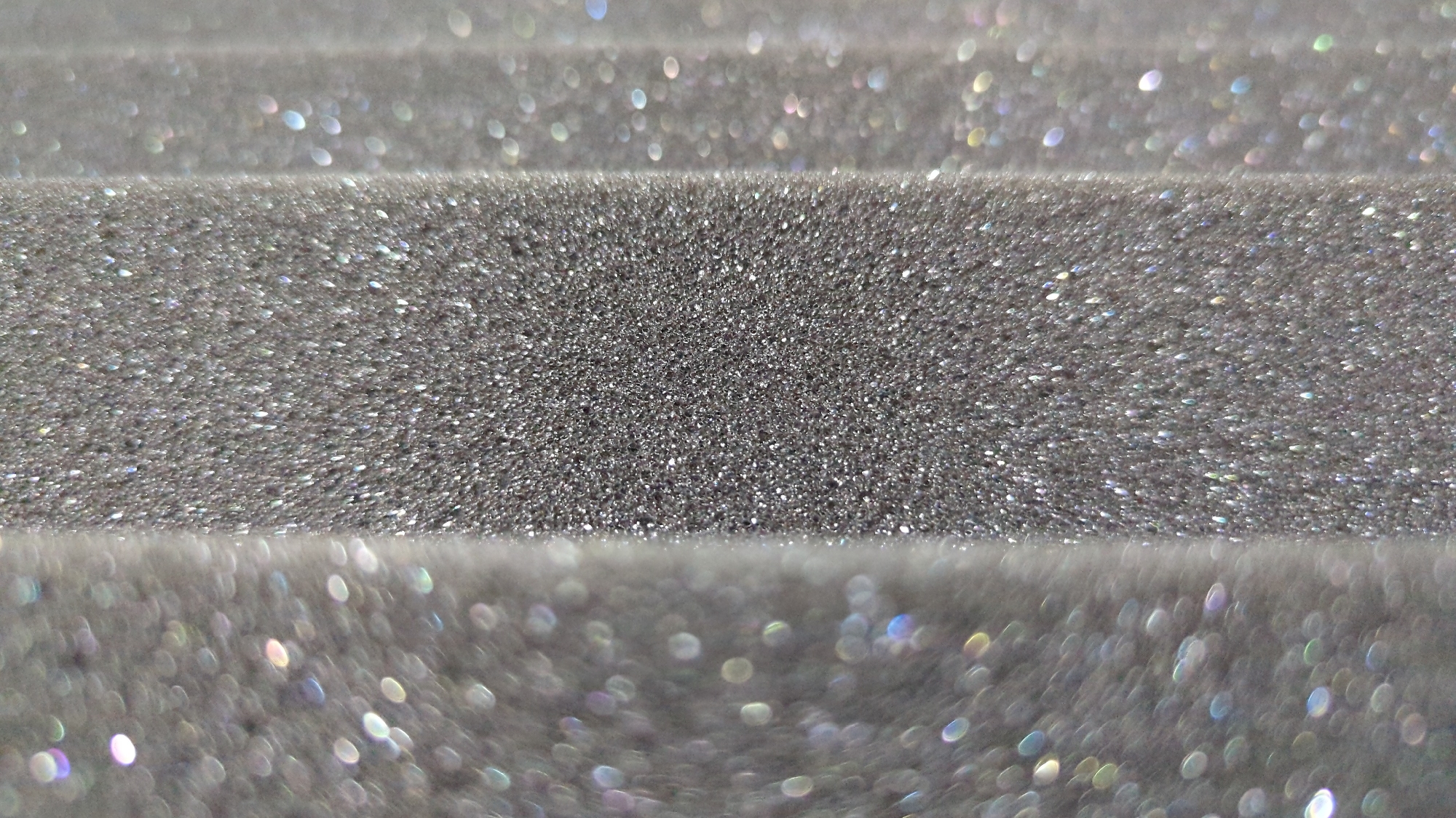 This is a close-up, abstract macro photograph of a textured, silver-gray surface. The surface appears to be a form of foam or sponge with a fine, granulated texture, possibly embedded with glitter or reflective particles. Vertical lines or divisions in the texture are visible, suggesting a layered or segmented material. The lighting creates numerous small, out-of-focus highlights that appear as bokeh circles and elongated shapes with iridescent colors, primarily white, blue, and pink. The image is devoid of any discernible human figures, animals, or recognizable objects from a wider environment. It is a purely textural and light-play study. There is no information within the image to suggest a location in Penjor, Indonesia.