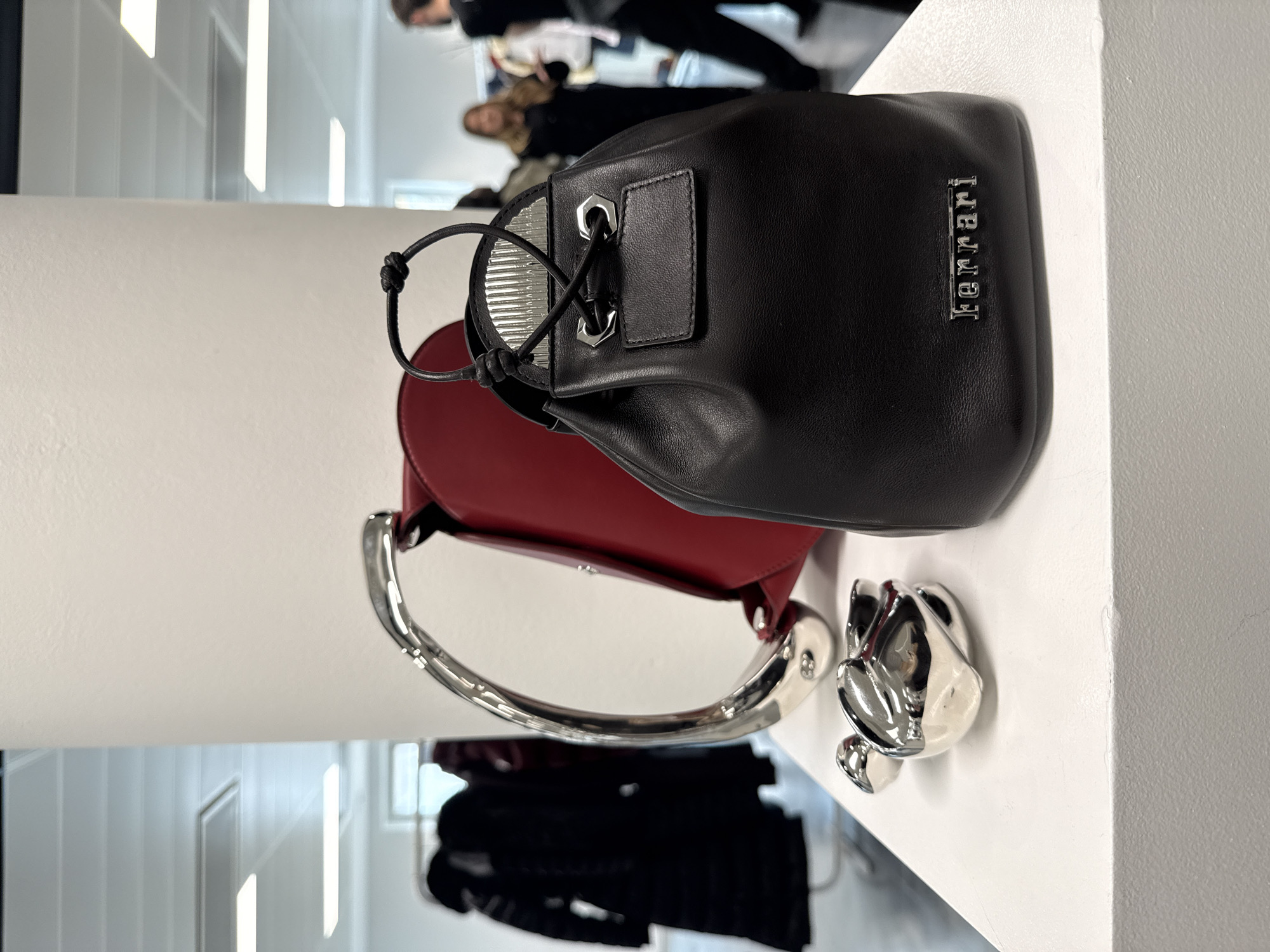 The photograph captures a collection of objects displayed on a white surface, likely a shelf or table, in what appears to be an interior setting. Dominating the foreground are two handbags. One is a black, bucket-style bag with a silver "Ferrari" logo vertically aligned on its side. The bag has a drawstring closure with silver eyelets. The other bag, primarily red, features a unique, curved silver handle. Next to the bags is a silver-colored, abstract figurine resembling a floral shape or a stylized animal.
In the blurred background, there are indistinct figures of people. They are positioned in a less defined area, indicating they are further away or intentionally out of focus. The interior setting features a white color scheme, suggested by the wall and the surface on which the items are placed. The lighting seems bright and even.
It's not possible to ascertain the location in London or the United Kingdom from the given information.