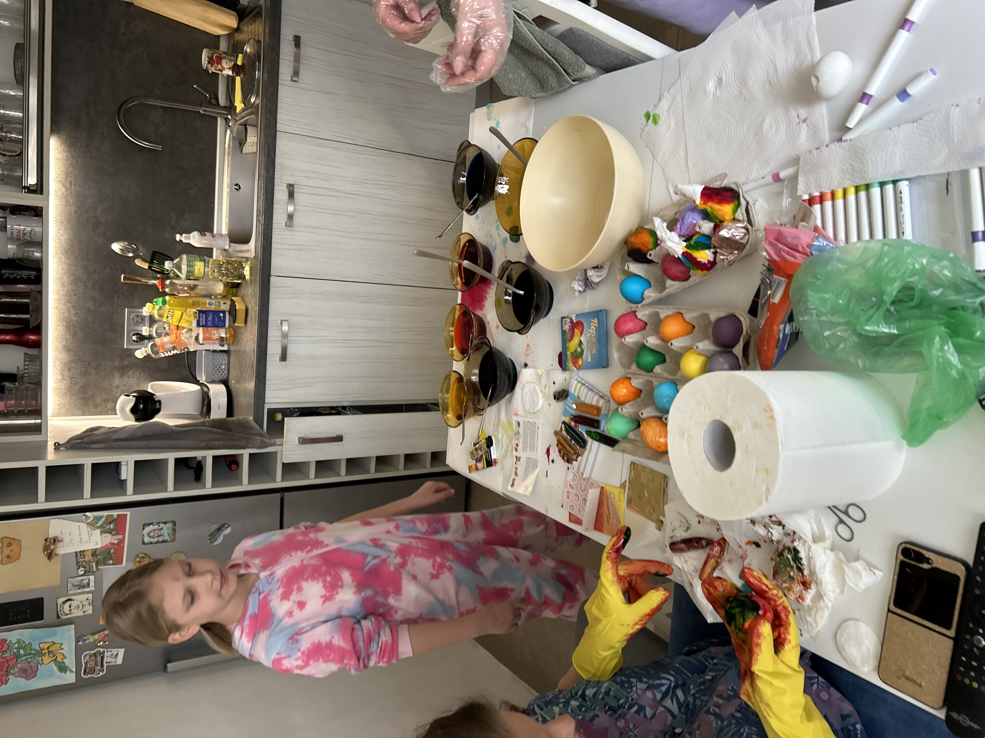The image depicts an indoor scene, likely a kitchen, where individuals are dyeing Easter eggs. A fair-skinned girl in a pink and white tie-dye shirt stands near a table. Two other people, possibly adults, are seated at the table, one wearing yellow gloves and the other clear plastic gloves. They appear to be actively involved in the egg dyeing process. The table is covered with various items including bowls containing dye, cartons of eggs, markers, a roll of paper towels, and a mobile phone. The environment suggests a domestic setting with kitchen cabinets, appliances, and decorative magnets on the refrigerator. There is no specific indication of Sofia, Bulgaria.