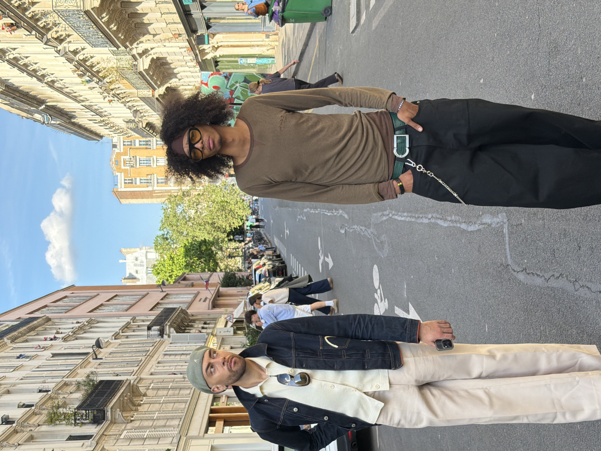 Two young men are standing in a street in Paris, France. The street is lined with buildings. The man on the left is wearing a dark-colored denim jacket, beige pants, a green hat, and a light-colored collared shirt with sunglasses hanging down. He is holding a cell phone in his hand. The man on the right is wearing a brown long-sleeved shirt, black pants, a green belt, and glasses. People are walking and standing in the background.