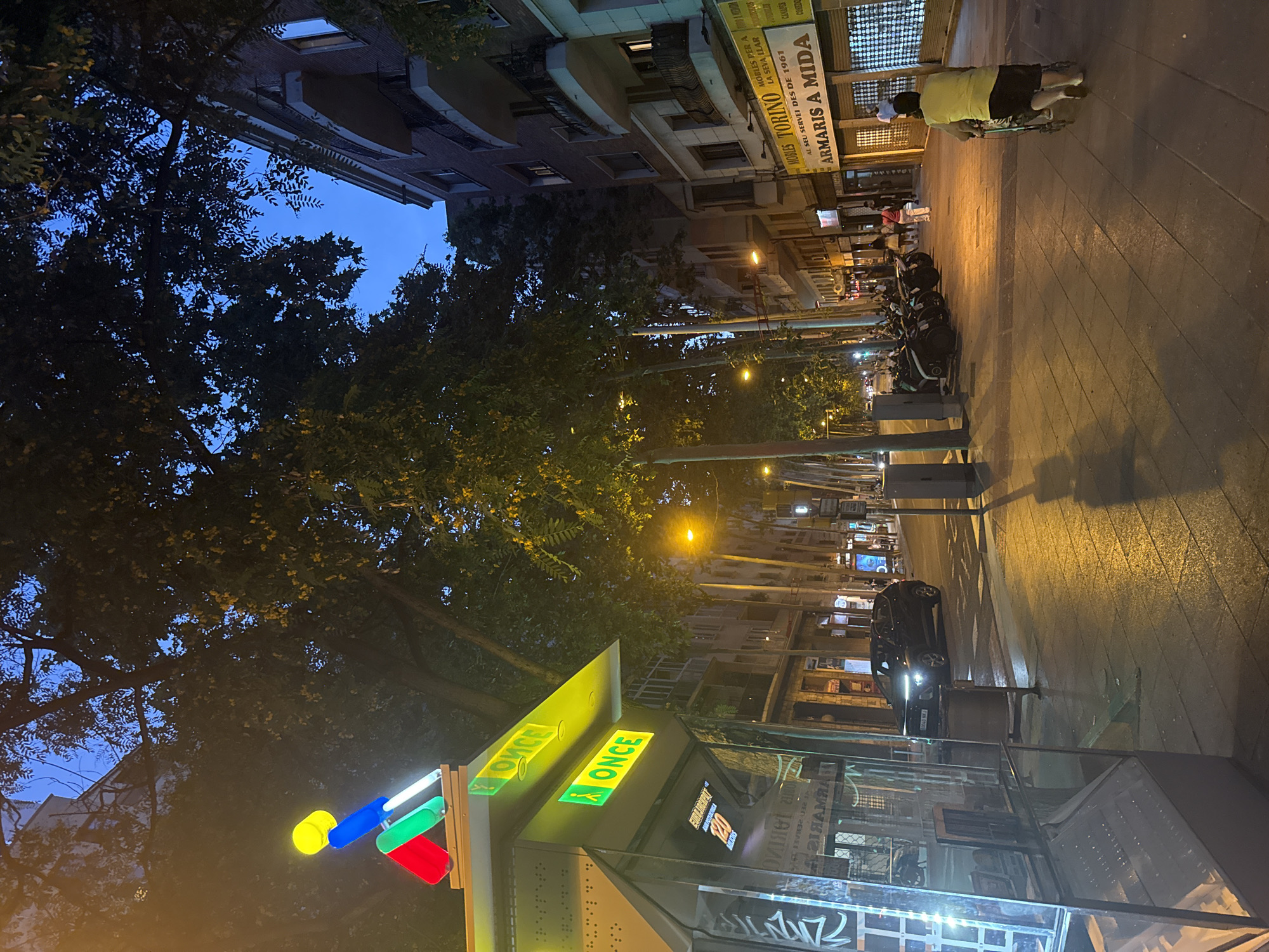 The image depicts a street scene in Barcelona, Spain, at dusk or early evening. The foreground features a glass-enclosed kiosk with prominent green "ONCE" signage, indicating a lottery and social services organization. Above the kiosk, a colorful abstract sculpture with red, green, and blue elements is illuminated. The street itself is paved with stone tiles, and lampposts cast a warm glow on the sidewalk and road.
Several motorcycles are parked in a row along the sidewalk, suggesting a busy urban area. A few pedestrians are visible: a woman pushing a stroller in the mid-ground and a person walking away from the camera further down the street. A black car is driving on the road in the middle ground.
The background is dominated by multi-story buildings, characteristic of Barcelona's urban architecture, with balconies and windows. Trees with dark foliage line the street, their leaves silhouetted against the deep blue twilight sky. Shops with illuminated signs are visible along the street, including one with the sign "ARMAMARIS A MIDA" and "MOBLES TORINO". The overall atmosphere is one of an active city transitioning from day to night.