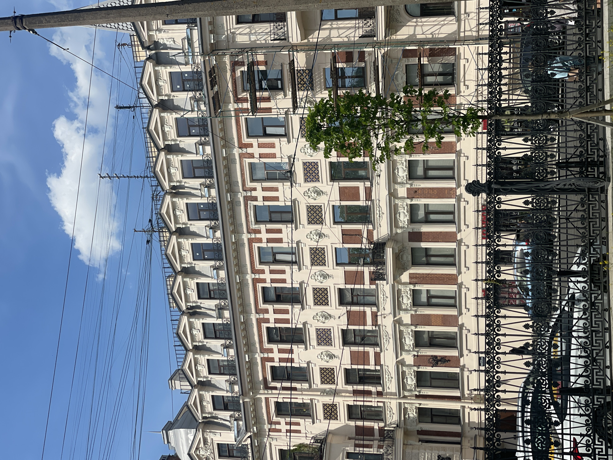 The image shows a view of a building facade in Saint Petersburg, Russia, from street level. The building is multiple stories high, with a decorative facade featuring a pattern of windows, brickwork, and ornate architectural details. A wrought-iron fence runs along the sidewalk in front of the building. A small tree is visible in front of the facade. Power lines extend across the sky above the building, set against a blue sky with a white cloud. The image is taken from a low angle, looking upward.