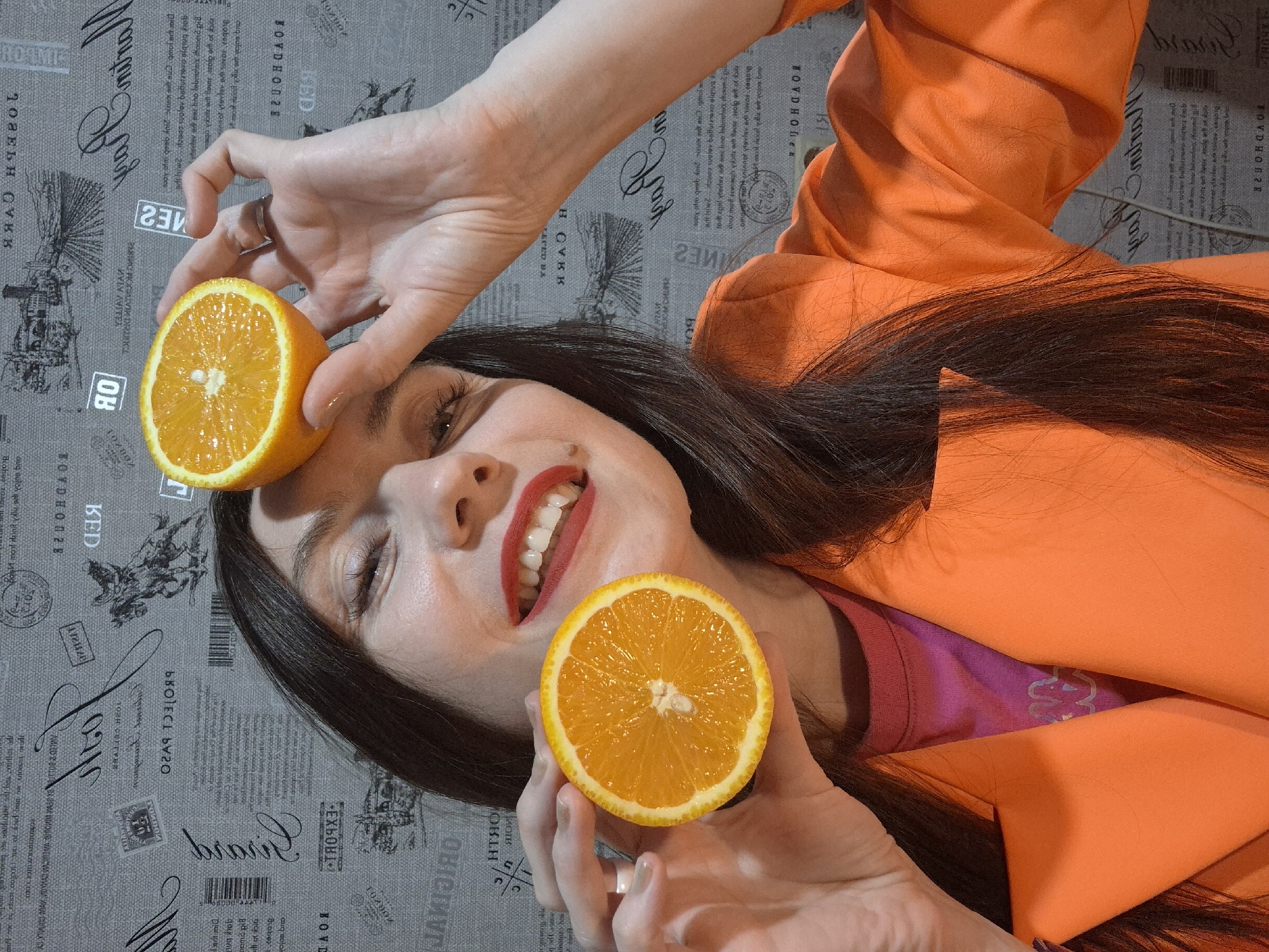 The image features a woman lying on her back, with her head oriented towards the bottom of the frame and feet towards the top. She is holding halves of an orange, one near her forehead and the other near her chin. She has dark hair and red lipstick, and is wearing an orange blazer over a pink top. The background is a gray patterned surface.
