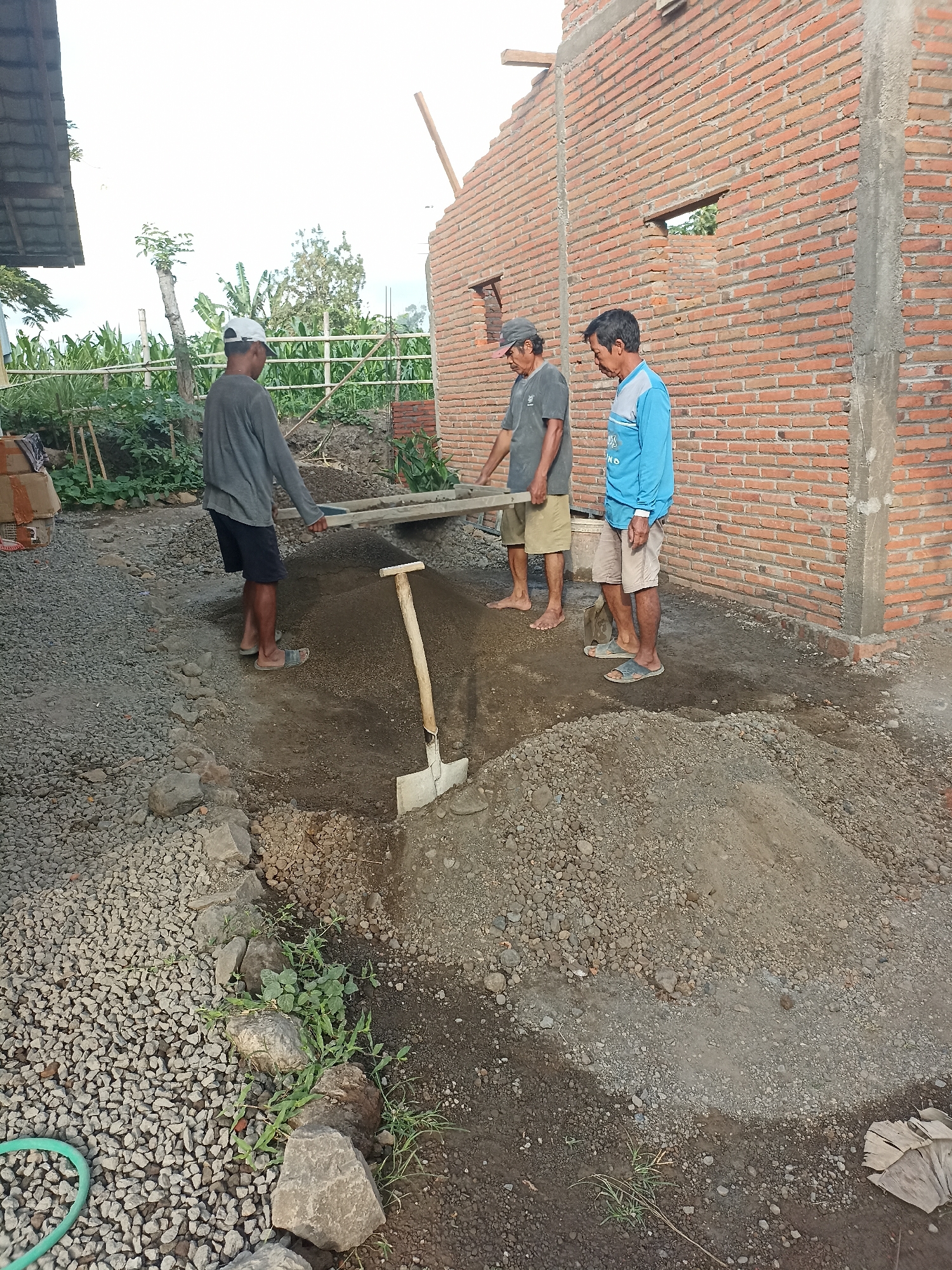 Three men are working outdoors on a construction site in Mojopanggung, Indonesia. The scene depicts an unfinished brick building with exposed mortar and unfinished walls. One man, wearing a gray long-sleeved shirt and dark shorts, is carrying a wooden plank. Another man, in a dark gray t-shirt and light brown shorts, is walking beside him. The third man, wearing a blue long-sleeved shirt and light brown shorts, stands near a pile of dirt. A shovel is partially buried in the dirt pile. The ground is a mix of gravel and dirt, with some scattered rocks and vegetation. The sky is overcast.