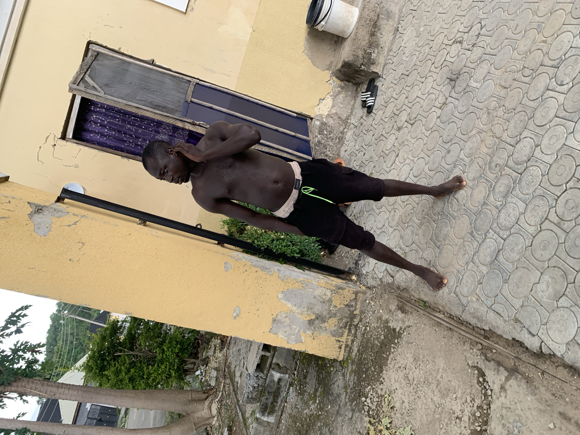 In an outdoor, urban environment with a backdrop of a pale yellow, weathered building, a dark-skinned man stands to the left of a partially open, weathered wooden door. He is shirtless, wearing black cargo shorts with a light-colored drawstring, and has his left hand touching his ear. The ground is paved with interlocking concrete tiles, and to the right of the man, a white bucket with a black lid rests on a raised concrete ledge. Beside the bucket are a pair of black and white Adidas slides. In the background, to the left of the building, are two palm trees, a dense green bush, and further back, a glimpse of other buildings and utility poles. The overall scene suggests a residential or commercial area in Abuja, Nigeria, with visible signs of aging infrastructure and common urban elements.
