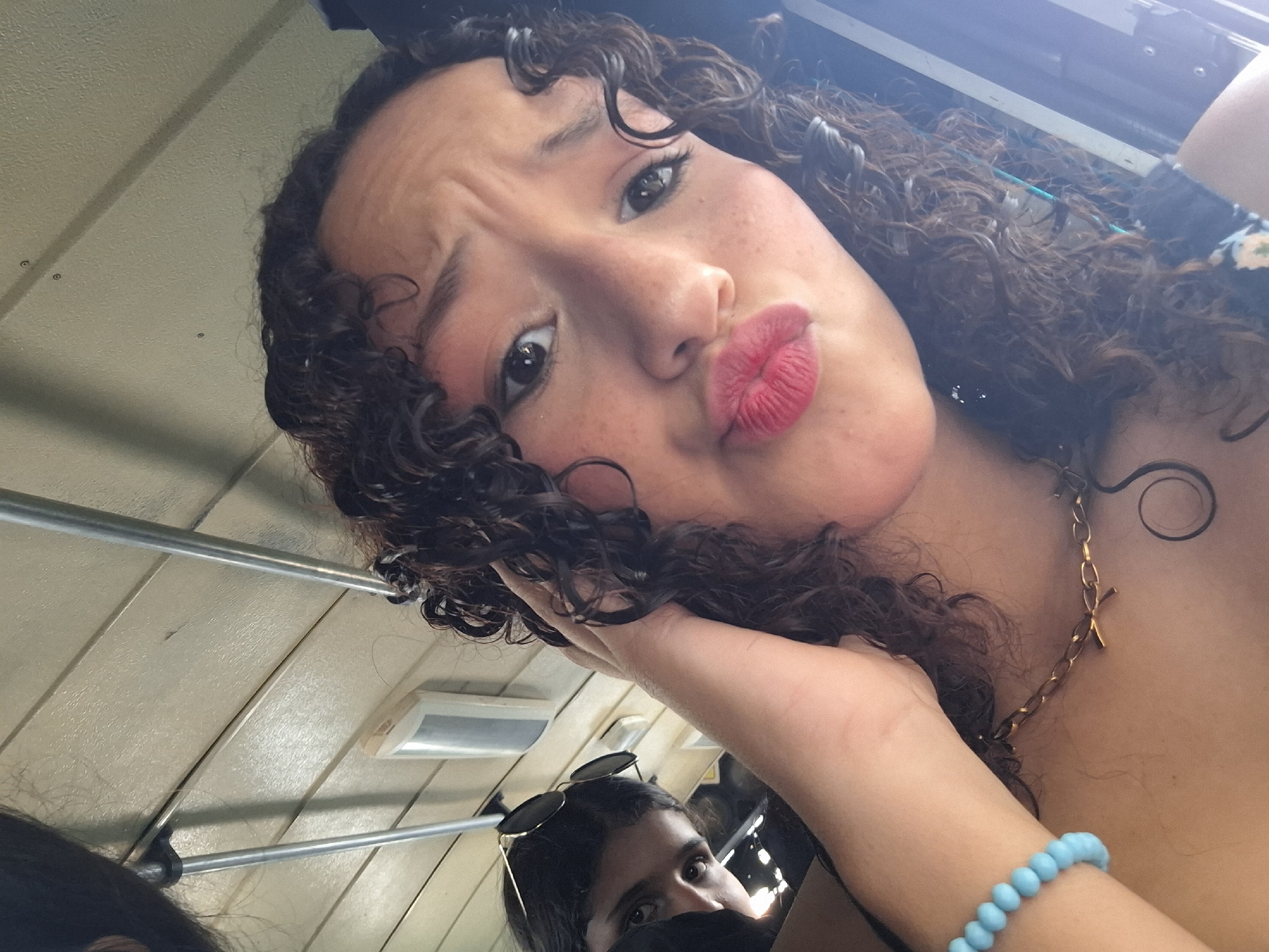 The image is a close-up, high-angle selfie of two young women, possibly friends, taken outdoors. The woman in the foreground has curly dark hair, light skin with freckles, and is puckering her lips, looking at the camera with a playful expression. She is wearing a gold necklace with a cross pendant and a light blue beaded bracelet. Her right hand is resting on her cheek, and her arm is visible.
The woman in the background is partially visible, wearing sunglasses perched on her head and a dark covering over her nose and mouth. Her eyes are looking towards the camera.
The setting appears to be inside a vehicle, possibly a bus or ferry, with light-colored wooden paneled walls and metal railings. The lighting suggests it's daytime, with a bright light source coming from above, casting shadows. There is no visible text in the image. The overall mood is casual and fun, typical of a selfie with friends.