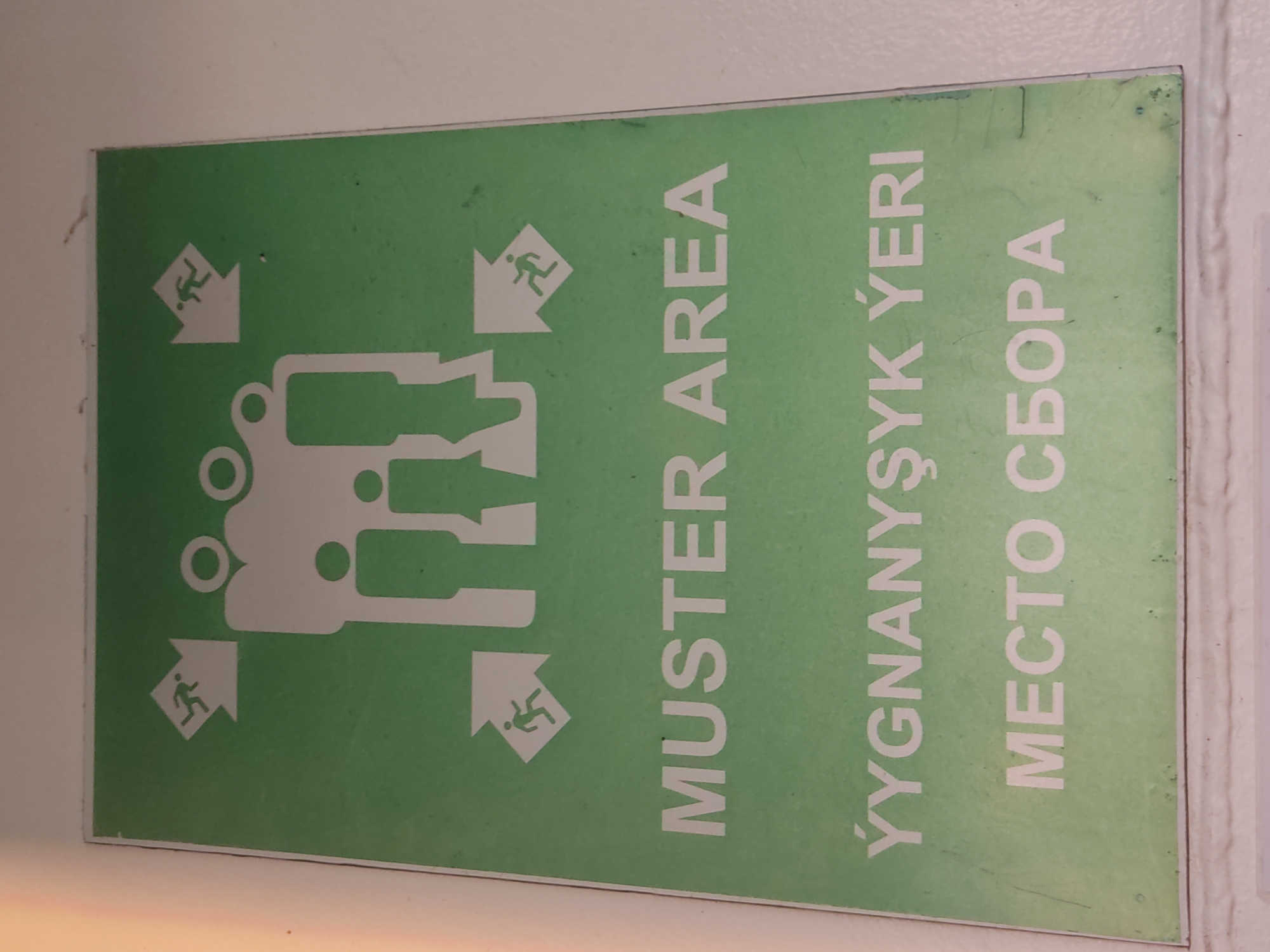 The image shows a green sign with white text and graphics. The sign is oriented vertically, with the text and graphics running from bottom to top. The graphics depict a stylized human figure running or walking, with arrows pointing towards a central gathering point represented by interlocking shapes. There are also smaller figures of people running into arrows, indicating an evacuation route.
The text on the sign is in three different languages. The leftmost text reads "MUSTER AREA". The middle text reads "YYGNANYŞYK ÝÉRI". The rightmost text reads "MECTO СБОРА". The sign appears to be an emergency evacuation or assembly point sign.
The setting is an indoor location, likely a public building or facility, indicated by the white wall against which the sign is mounted. The lighting appears to be artificial. There are no discernible clues about the time of day or weather. The overall impression is one of clear signage for a safety procedure. The sign is slightly weathered, with some scuffs and marks, suggesting it has been in place for some time.