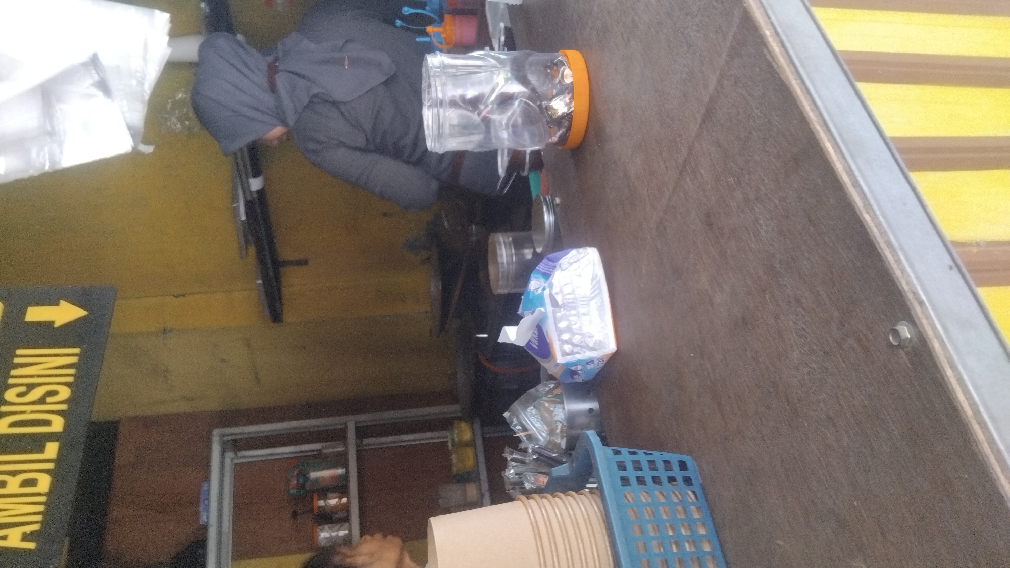 The image depicts an indoor scene, likely a food stall or small restaurant, with a focus on the wooden countertop in the foreground. To the left, a blue plastic basket holds a stack of beige paper cups. Behind the cups, a metal shelf unit contains several jars, some filled with condiments or food items. A person wearing a grey headscarf is partially visible on the left, facing away from the camera. In the background, to the right, another person wearing a grey headscarf and a grey top is seen from the back, appearing to be cooking or preparing food. They are standing in front of a yellow wall. A clear plastic jar filled with small, metallic objects and an orange lid sits on the countertop. A white and blue packaged item, possibly a food product or tissues, is also on the counter. The overall environment appears functional and somewhat cluttered, typical of a small business. There is a sign with yellow text on a black background that reads "AMBIL DISINI" with a yellow arrow pointing down. The presence of this sign suggests a designated pick-up area. The location is not specified within the image itself, and therefore its connection to Proyonangg, Indonesia, cannot be confirmed from the visual information.