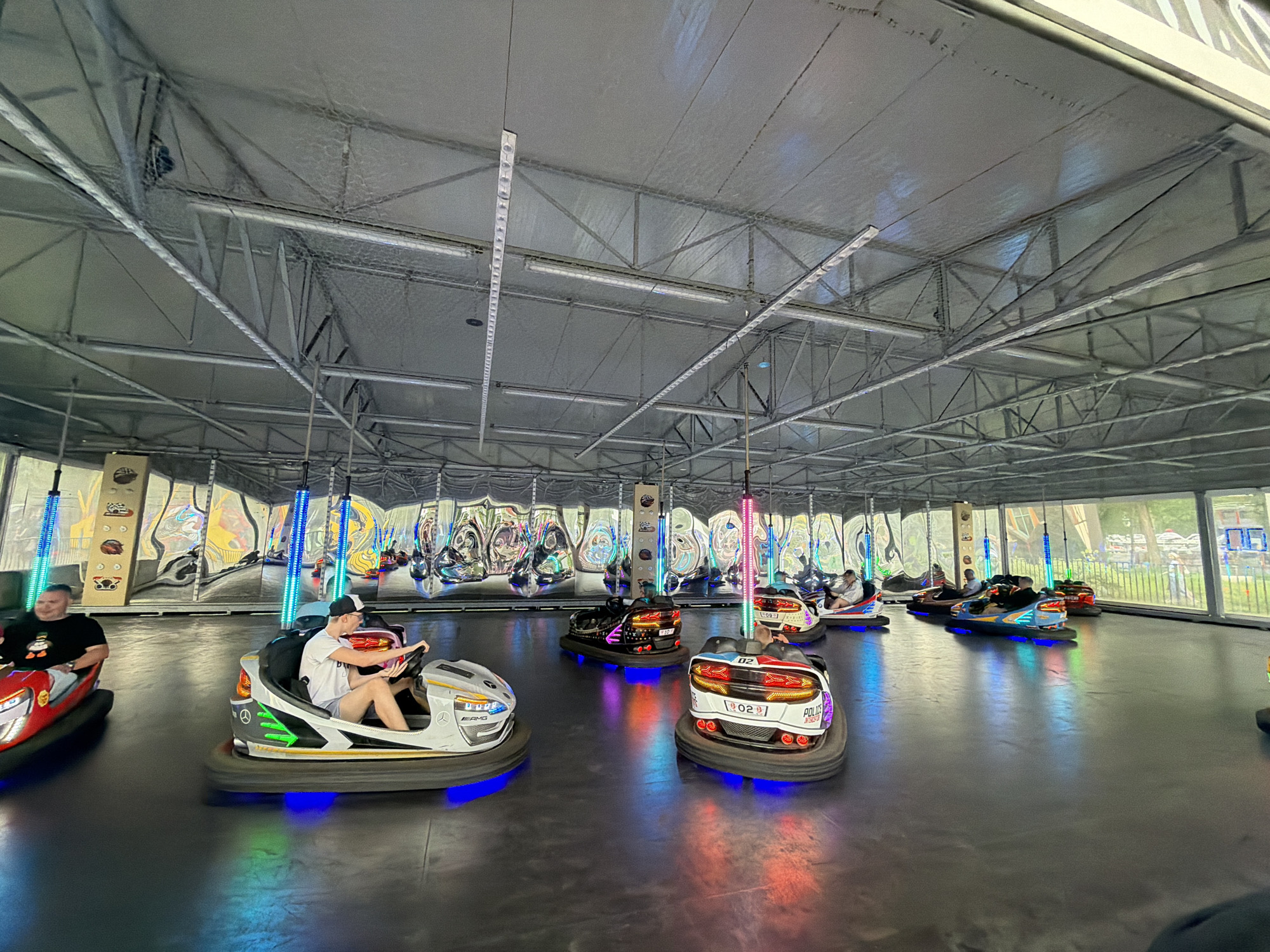 The image depicts an indoor bumper car arena with a group of people enjoying the ride. The arena has a metal truss ceiling with numerous lights and wires. The floor is dark and reflective, illuminated by the colorful lights of the bumper cars. Several bumper cars are in motion, with drivers operating them. One bumper car in the foreground is white with silver accents and "AMG" branding, occupied by a person wearing a white t-shirt and shorts, with a black and white baseball cap. To the left of this car, another bumper car, red with silver accents, is occupied by a person in a black t-shirt with a cartoon character. In the background, more bumper cars and people are visible, along with decorative panels on the wall featuring various abstract and figurative designs. The overall atmosphere suggests a recreational activity in a well-lit, enclosed entertainment venue.