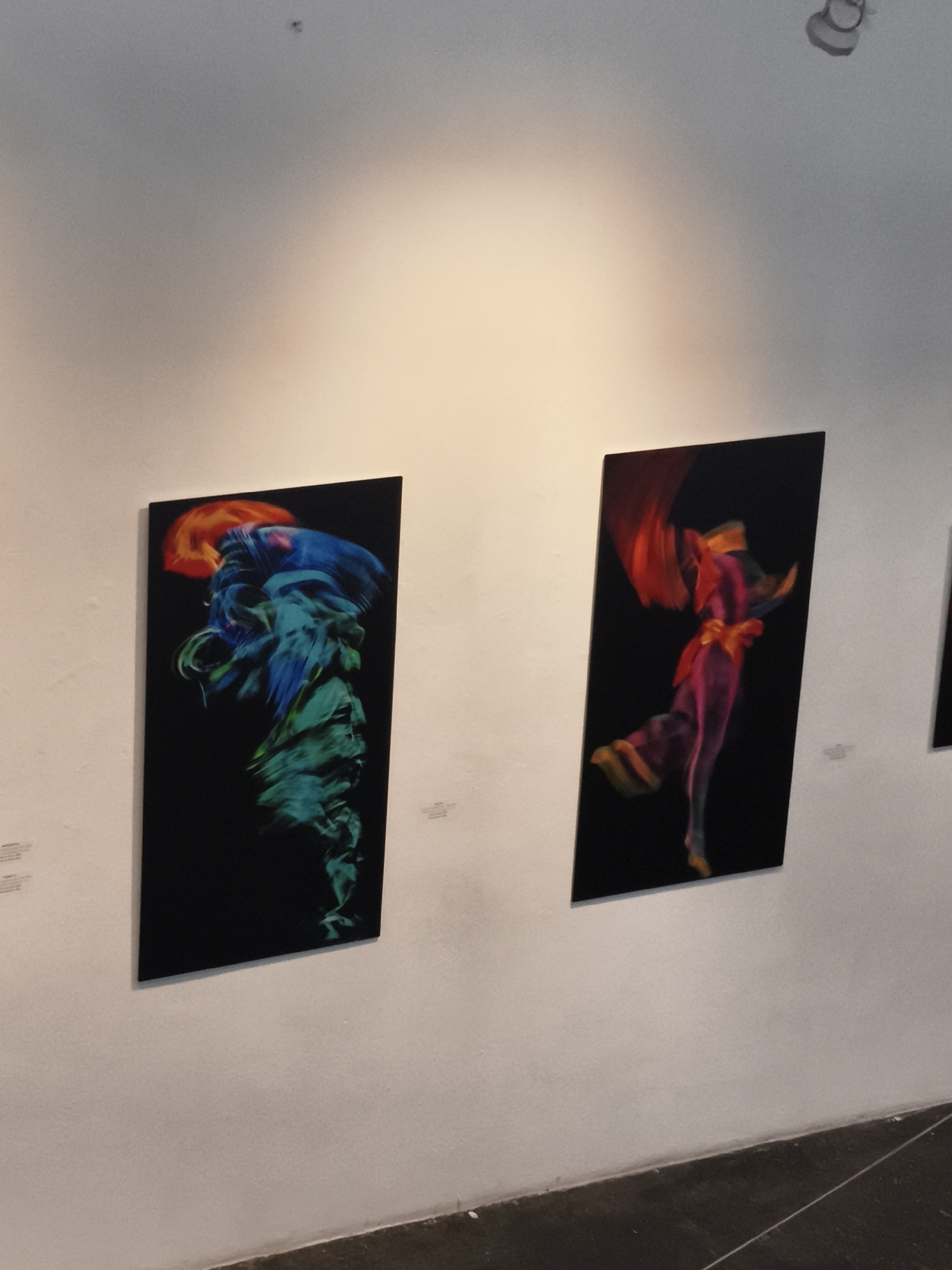 The image shows two pieces of artwork displayed on a white wall, likely in an art gallery in the city of Querétaro, Mexico.
The artwork on the left depicts an abstract, multi-colored figure in blue, green, and orange against a black background. The artwork on the right shows a similarly abstract human figure in motion, depicted with red, pink, and orange hues, also set against a black background.
Both artworks are framed in black. There are unreadable labels or descriptions on the wall next to the artworks. The environment is modern and minimalist. A ceiling light is visible above the artwork.