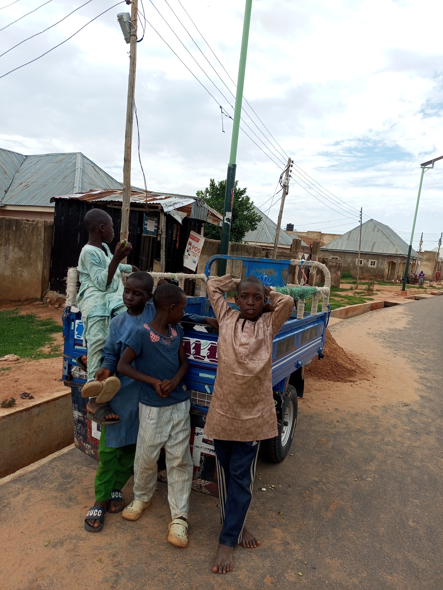 In Tempure, Nigeria, four young boys are gathered near a blue three-wheeled truck parked on a dusty roadside. One boy stands on the truck bed, holding a green fruit and wearing a light blue tunic and pants. Two other boys stand beside the truck; one in a dark blue t-shirt and striped pants, the other in a blue tunic and green pants, holding a pair of sandals. A fourth boy, wearing a tan tunic and dark pants, stands in front of the truck with his hands behind his head, looking at the camera. The truck appears to be loaded with sand. Buildings with corrugated metal roofs are visible in the background, along with utility poles and power lines, suggesting a residential area. The sky is overcast.