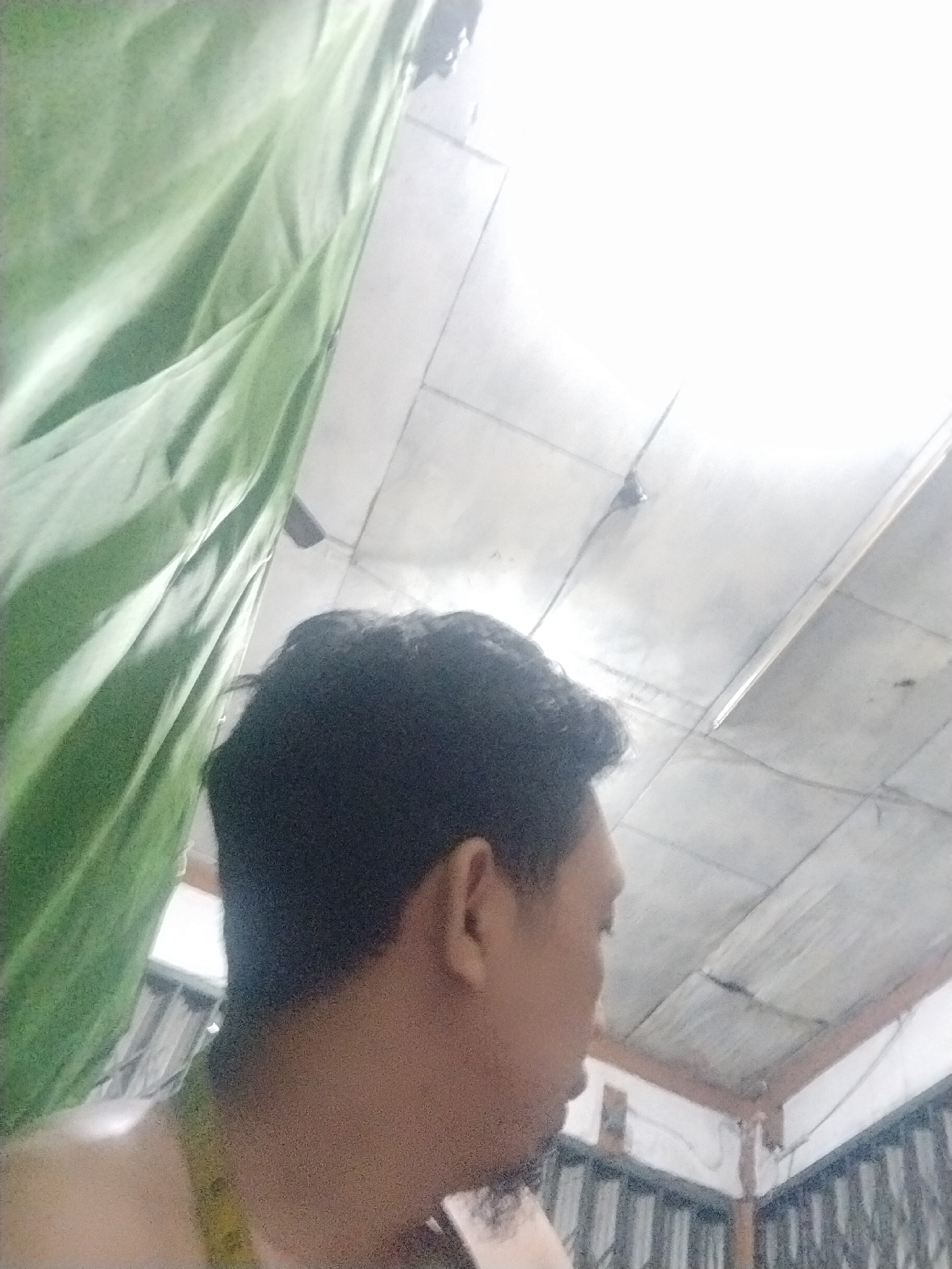 A man is shown from the shoulder up, in profile, facing left. He has dark hair and is wearing a light-colored garment around his neck. Behind him, a green curtain or fabric is visible on the left side of the image. The background consists of a ceiling with visible paneling and some lighting fixtures. The image appears to be taken indoors. The specific location in the city of Unknown, Indonesia cannot be determined from the visual information provided.