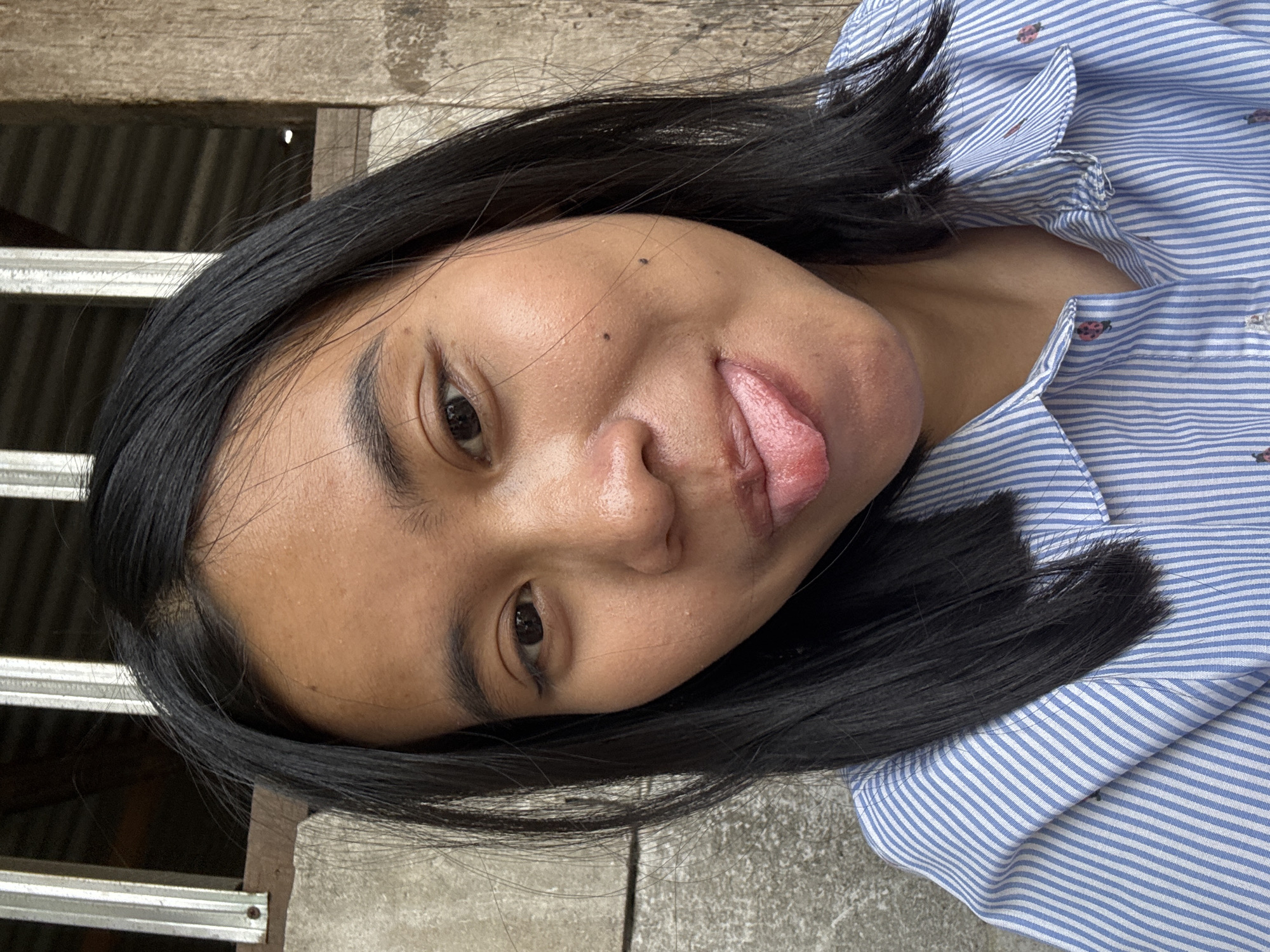 In East Jakarta, Indonesia, a young woman with dark hair and brown eyes is captured in a close-up selfie. She is wearing a blue and white striped button-down shirt with small embroidered ladybugs. Her tongue is extended, and she is looking towards the camera. The background consists of a weathered wooden wall and a concrete surface. The lighting suggests it is daytime.