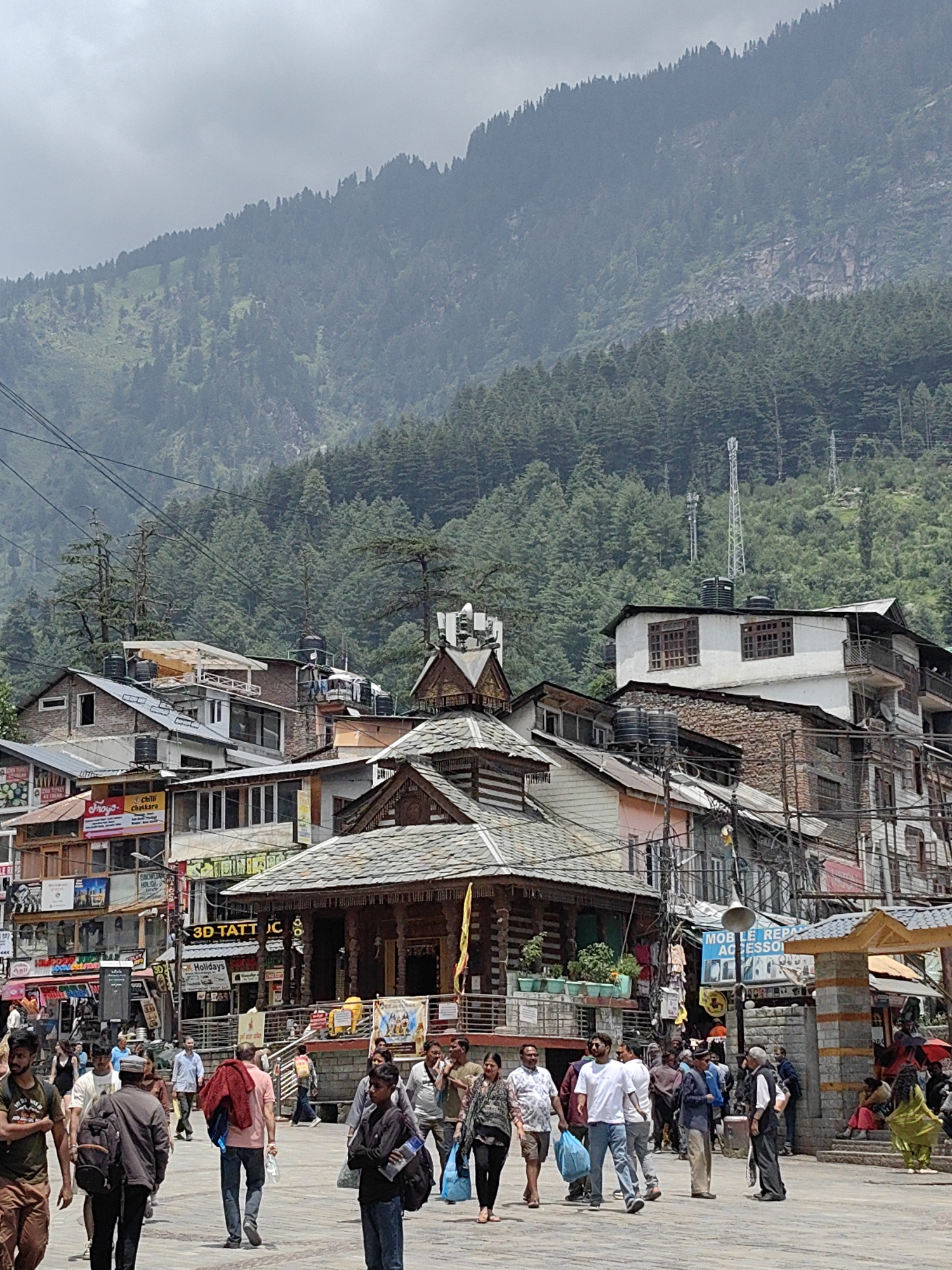 This image depicts a street scene in Manali, India, with a focus on the architecture and natural surroundings. In the foreground, a paved street is crowded with people walking in various directions. They are dressed in casual clothing, with some carrying bags or wearing backpacks. The street is lined with multi-story buildings, predominantly made of stone and wood, with tiled roofs. Many of these buildings appear to be shops or businesses, indicated by signs and displays. Some structures have balconies and multiple windows.
Behind the buildings, a dense forest of pine trees covers the slopes of a mountain. The mountain itself is steep and rocky, with the treeline reaching high up its face. The sky is overcast, suggesting a cloudy or potentially rainy day. A traditional wooden temple structure with a pagoda-style roof is visible in the middle ground, adding to the cultural ambiance of the scene. Electrical wires and poles are strung across the sky, typical of an urban or semi-urban environment. The overall impression is a bustling marketplace nestled within a scenic mountainous landscape.