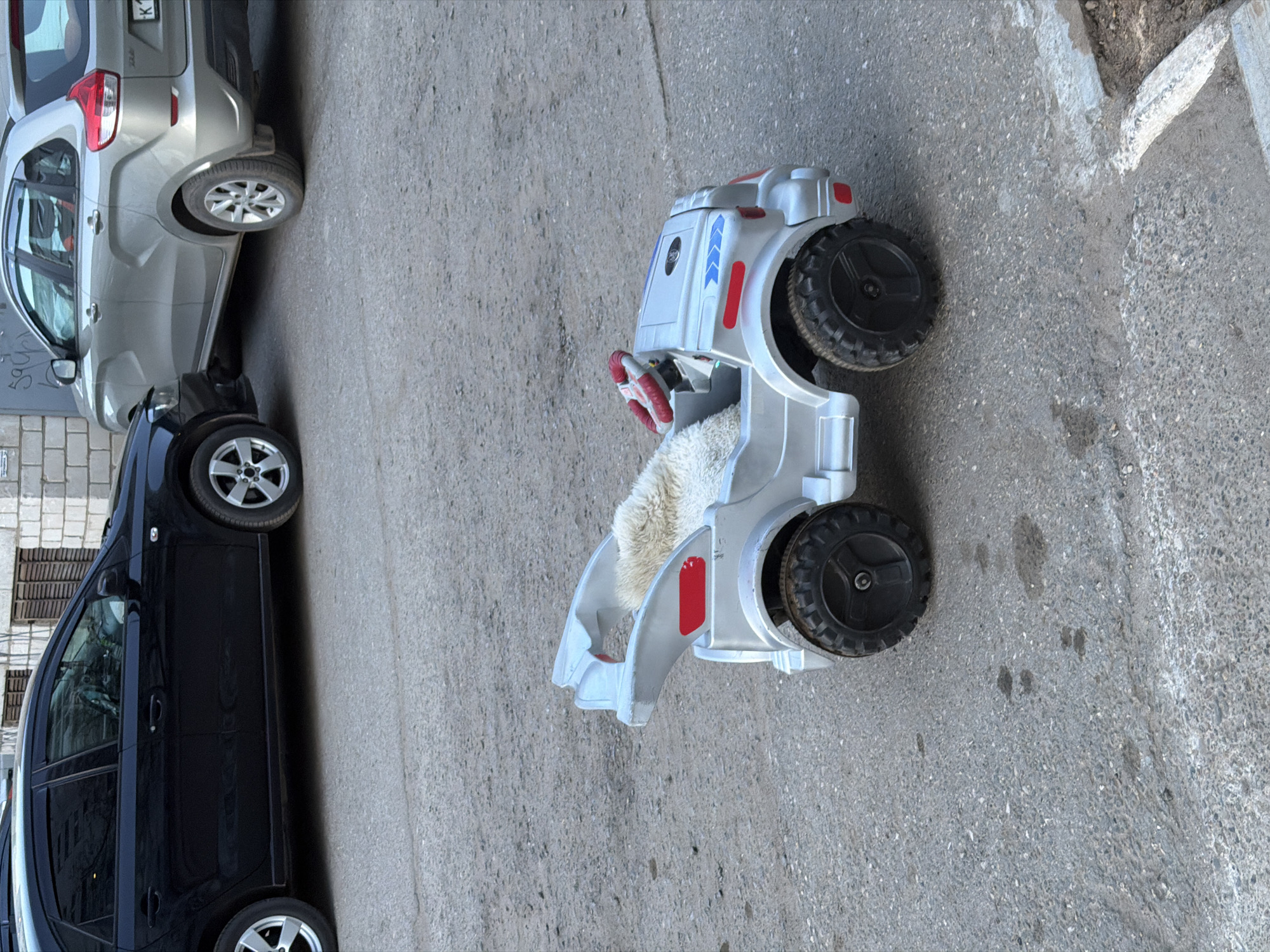 The image shows a street scene in what appears to be the city of Chaikovsky, Russia, evidenced by the vehicle license plates. A silver SUV-type vehicle is parked partially out of frame on the left, next to a black sedan. A children's toy car, white with red accents, is visible in the center of the frame. The toy car features oversized black wheels. The vehicles are parked on an asphalt surface adjacent to a concrete curb. The surrounding environment appears to be a residential area with a building partially visible in the background. No people are visible.