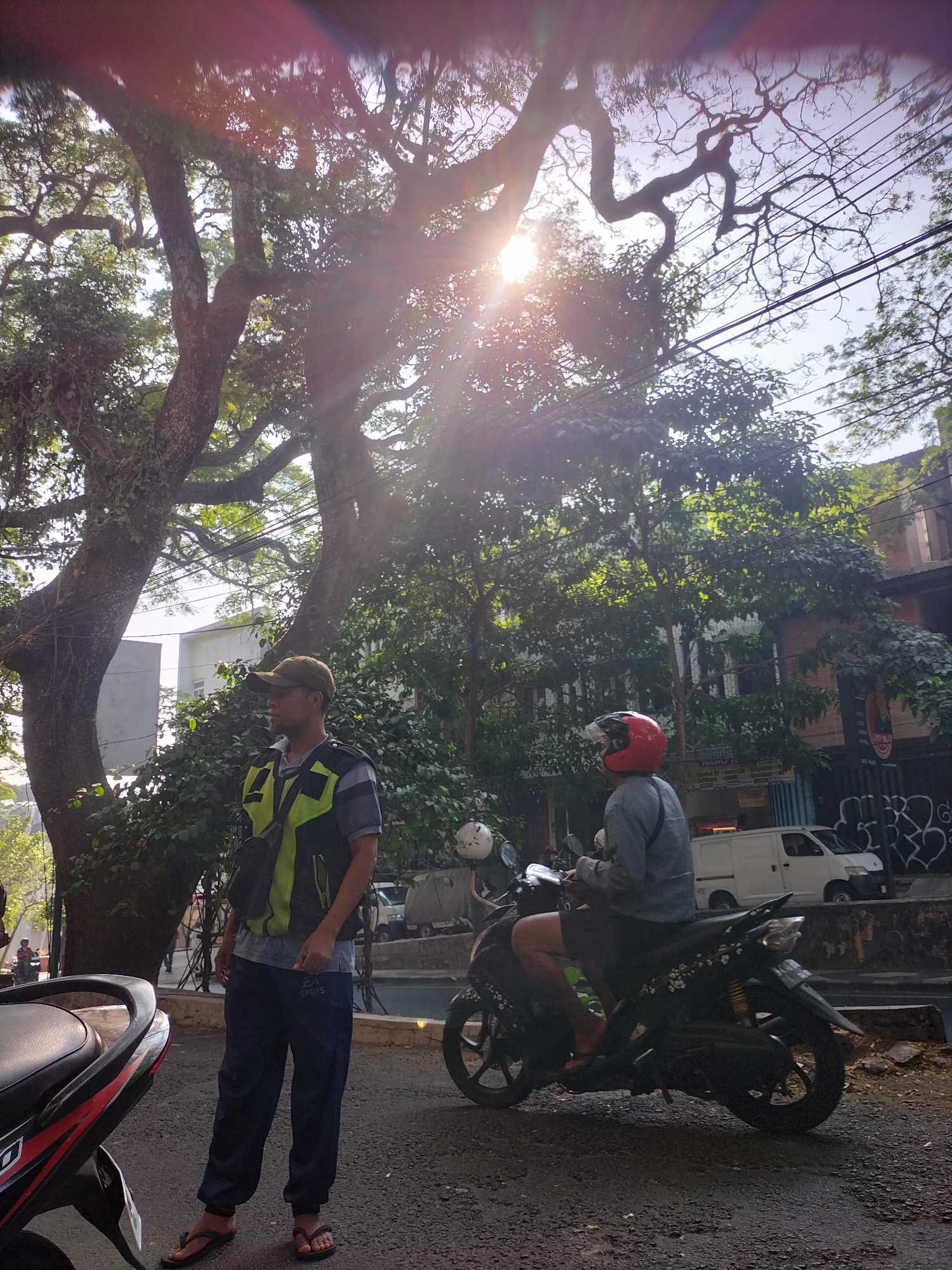 The image depicts an outdoor urban scene in Malang, Indonesia, characterized by bright sunlight filtering through large trees with dense foliage. In the foreground, a man stands on the right side, wearing a dark baseball cap, a neon yellow and grey vest over a grey shirt, and dark blue pants. He is looking to his left. To his right, a person wearing a red helmet and a grey long-sleeved shirt is seated on a dark-colored motorcycle, facing away from the camera. The motorcycle has a license plate.
In the background, several buildings are visible, including a light-colored building with windows and a darker building with graffiti on its lower section. A white van is parked in front of one of the buildings. The ground is paved with asphalt and gravel. The overall atmosphere is sunny and appears to be a typical day in an Indonesian city. The image captures a moment of everyday life, with individuals engaged in their activities amidst the urban environment.