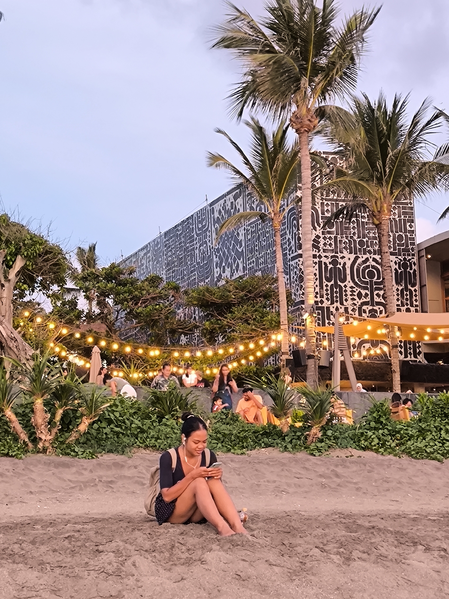 The image depicts a beach scene, likely in North Kuta, Indonesia, during the early evening. In the foreground, a young woman sits on the sand, looking at her phone with earphones in, and wearing a black top, shorts and a backpack. Behind her, a raised area hosts a gathering of people seated and enjoying the atmosphere. A string of lights is strung across the area, contributing to a warm ambiance. Tall palm trees punctuate the landscape, while a building with a distinctive black and white patterned facade stands in the background. The overall scene conveys a relaxed, beachside atmosphere.
