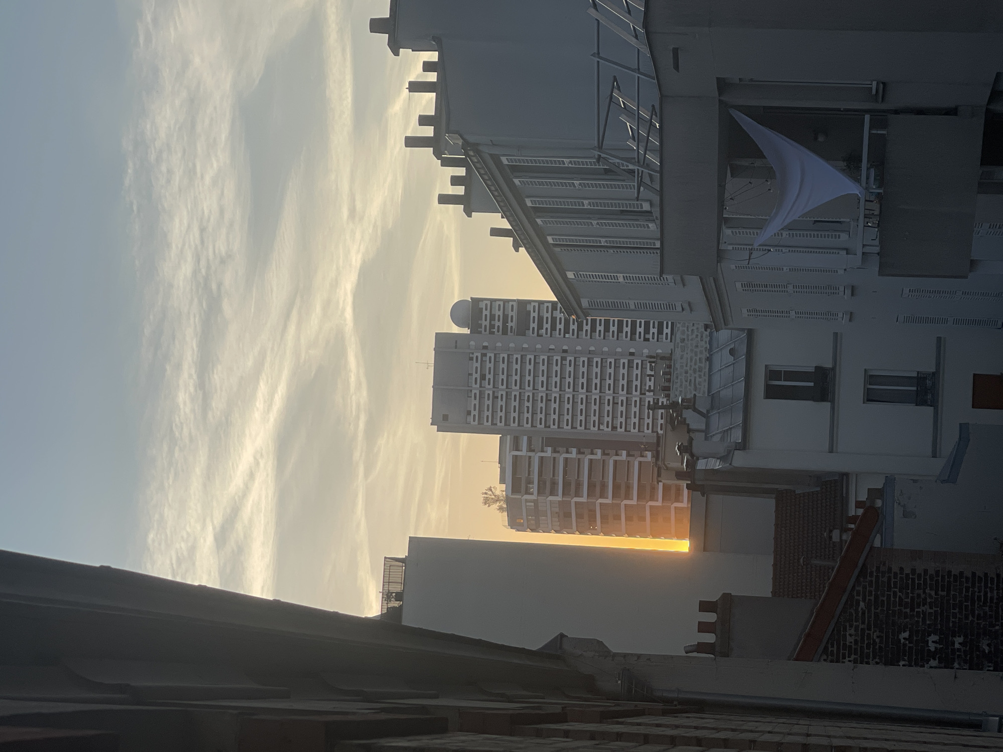 This is a vertical, eye-level, medium shot of a cityscape in Paris, France, taken during daylight, possibly at sunrise or sunset. The sky is a pale blue with wispy white clouds, illuminated by a warm, orange glow from the sun. In the foreground on the left, a dark, angled building with brick detail is partially visible, obscuring the view. The mid-ground is dominated by several tall buildings. On the far left, a slender, light-colored skyscraper stands tall. To its right, a lighter, multi-story building with repeating horizontal bands of windows and balconies is visible. Further right, a taller, white building with a distinctive rounded structure on its roof is prominent, its facade filled with numerous rectangular windows. In the foreground right, a light gray building with a flat roof and many closed shutters lines its windows. A balcony on this building features a large, white, triangular piece of fabric hanging down, possibly a sheet or a flag, suspended by cords. The scene is filled with architectural elements, including chimneys and rooflines, suggesting an urban environment. No people or animals are visible in the image. The overall atmosphere is calm and serene, bathed in the soft light of the sun.