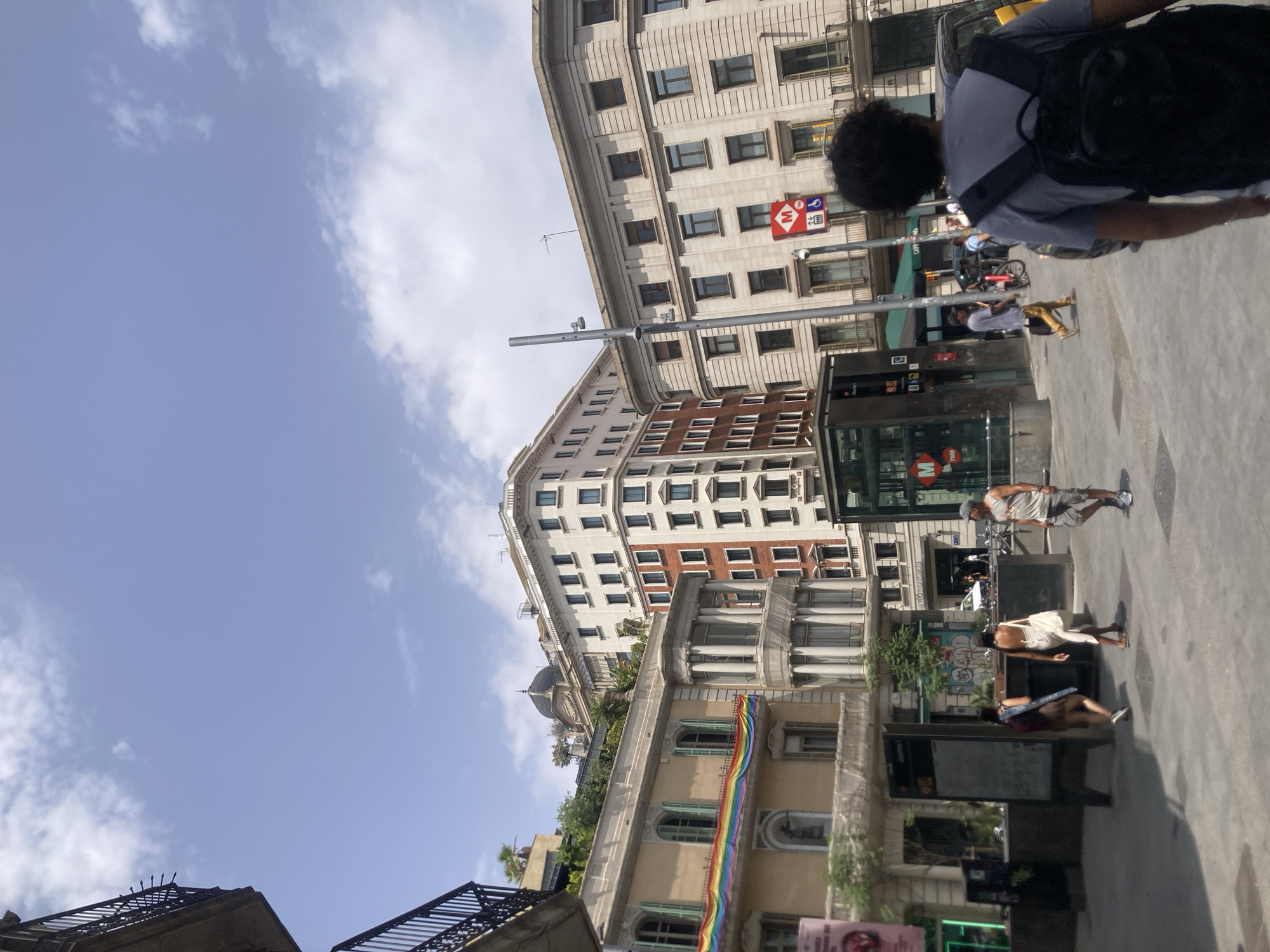 The image depicts a street scene in Barcelona, Spain, likely a commercial or tourist area, on a sunny day with scattered clouds. In the foreground, a person with dark, curly hair and a gray t-shirt with a black backpack is walking away from the camera, partially obscuring a yellow vehicle. The street is paved, and there are various buildings lining the street.
To the left, a building with a light yellow facade features green shutters and a rainbow flag draped across its upper story. Further to the left, a balcony with a black railing extends from a building.
In the middle ground, a modern glass-enclosed metro entrance with an "M" logo is visible. A red sign with an "M" symbol and a wheelchair accessible icon is also present on a pole. Several people are walking in different directions. Two women are walking together towards the left, one with a backpack and the other in a white dress. A man in a gray tank top and shorts is walking towards the right. Another man in a light blue shirt and khaki pants is walking to the left.
The background features a series of multi-story buildings with a mix of architectural styles. One building is clad in red brick, while others are light-colored with numerous windows and balconies. The sky above is blue with white clouds. The overall impression is of a bustling urban environment in Barcelona.