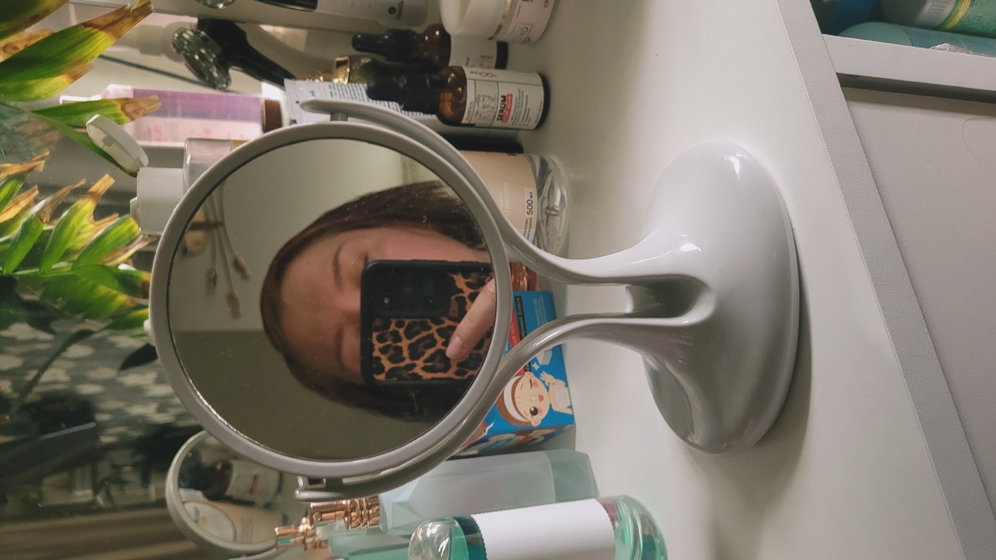 A person is taking a selfie in a mirror. They are holding a smartphone with a leopard-print case. The mirror is on a vanity with various beauty products and a plant. The overall scene is cluttered, suggesting a personal space like a bathroom or bedroom. The lighting is soft, indicating indoor illumination. No specific time of day or weather conditions can be inferred. Some visible text includes "SERUM" on a brown bottle and possibly labels on other products, but they are too blurry to read.