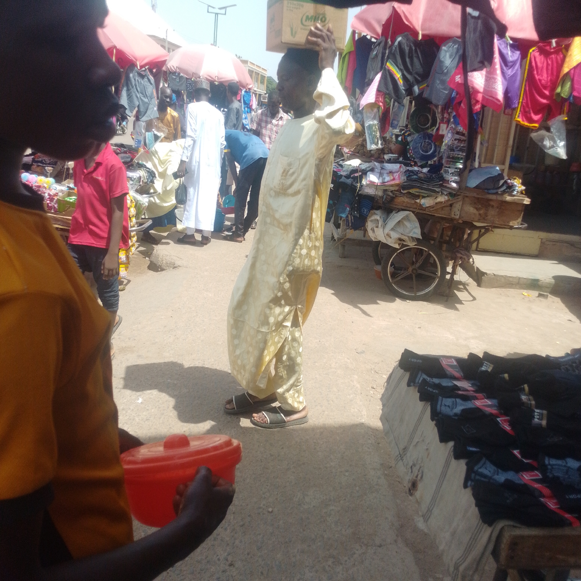 In Arawa, Nigeria, on a dusty street, a young boy in a bright yellow shirt holds a red plastic bowl. To his left, a boy in a red t-shirt stands near a market stall. Further back, a man in a light-colored, patterned robe carries a cardboard box on his head, walking amidst a bustling marketplace. The scene is filled with various stalls displaying clothing, with umbrellas providing shade overhead. A wooden cart laden with goods is visible in the background, suggesting commercial activity. The environment is outdoors, with a paved ground and the bright sunlight casting shadows.