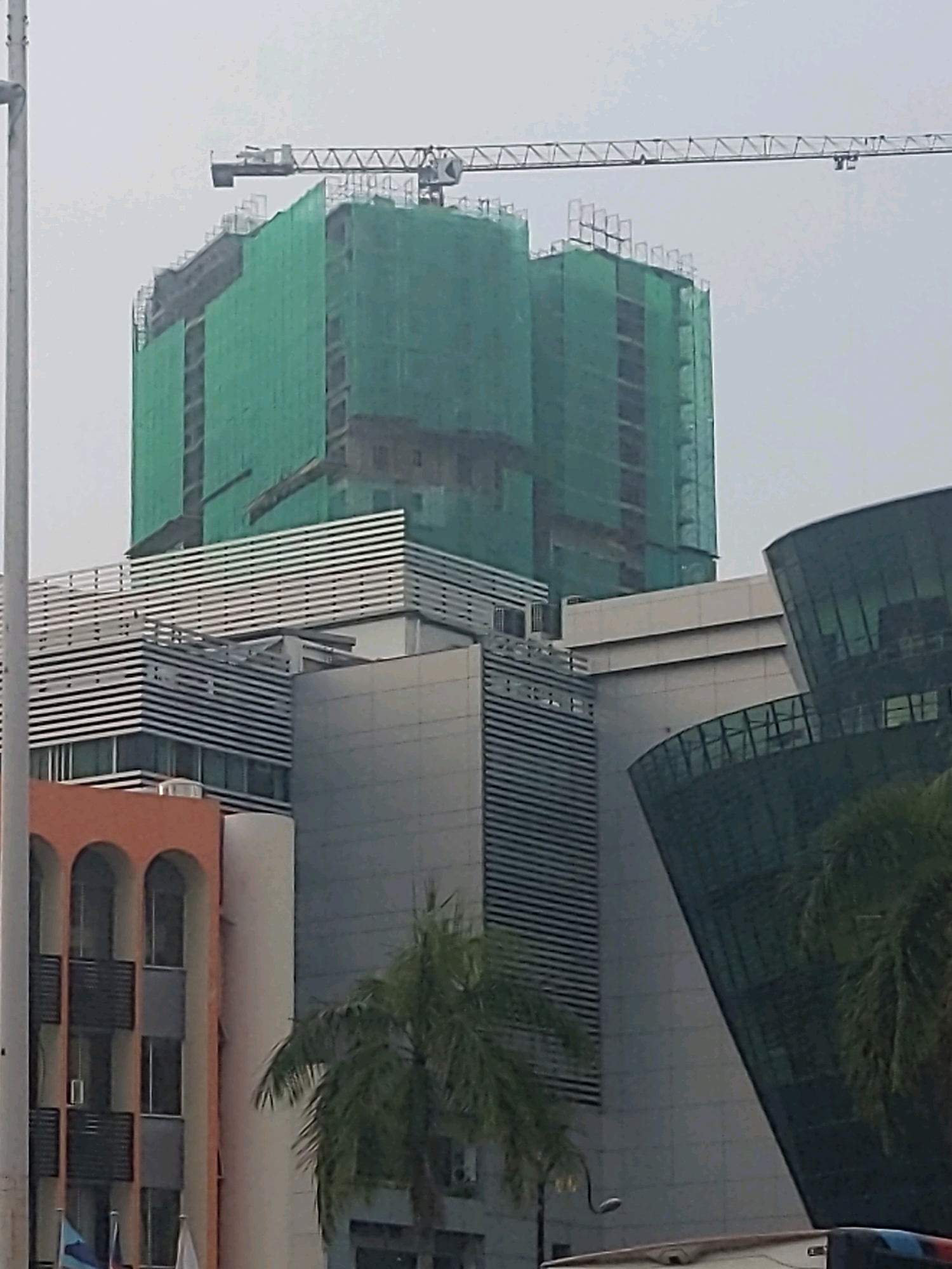 In Kota Kinabalu, Malaysia, a cloudy sky looms over a construction site featuring a large building covered in green netting, indicating ongoing construction. A tall crane extends over the structure, with its arm positioned horizontally. In the foreground, several modern buildings are visible. To the left, a building with orange arches and dark windows is partially obscured. To the right, a building with a curved, dark glass facade reflects the overcast sky. Between these structures, a palm tree adds a touch of greenery to the urban landscape. The image captures a typical scene of urban development in the city.