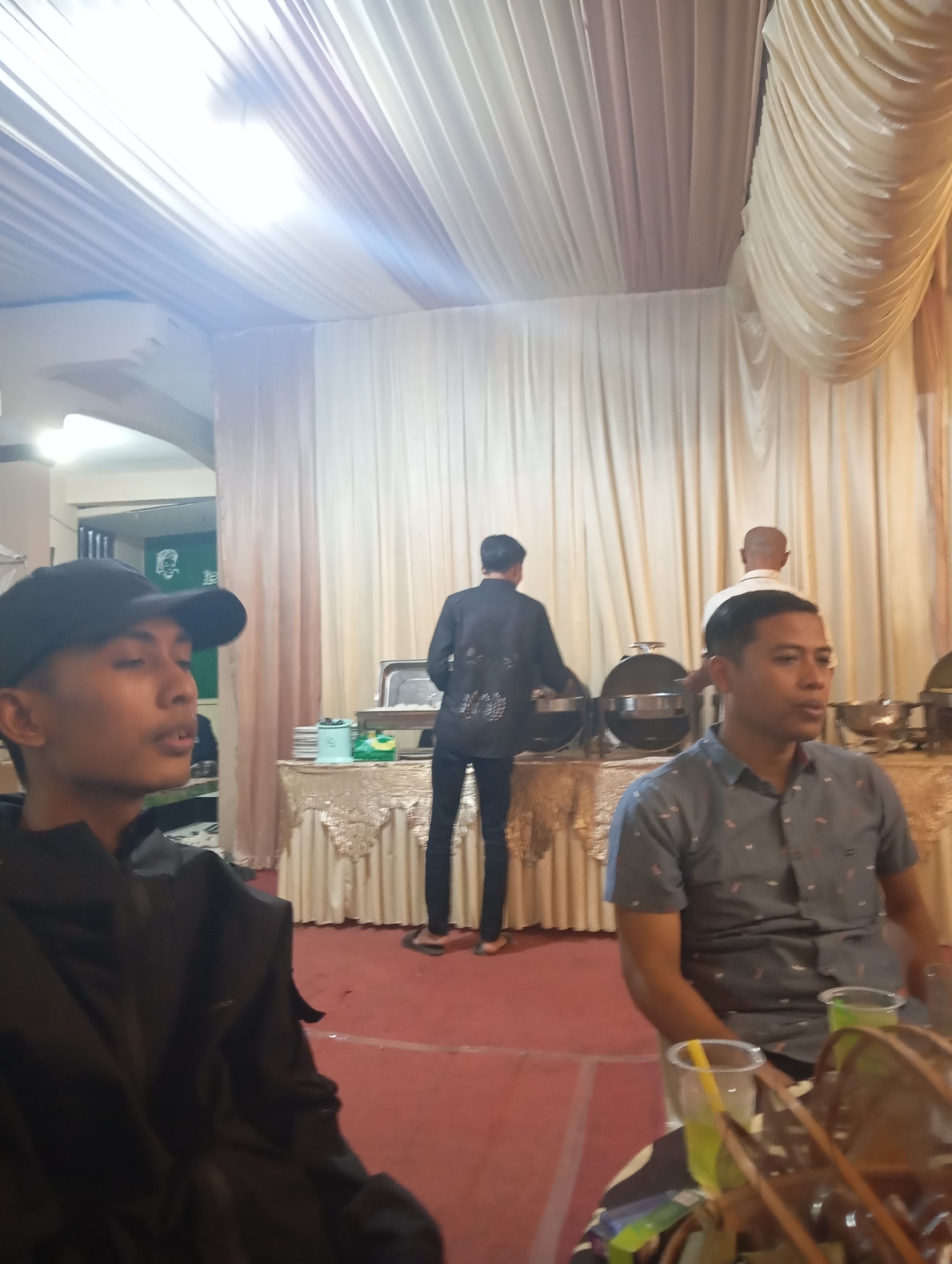 The image depicts an indoor event, likely a reception or gathering, with a prominent red carpet covering the floor. The space is decorated with draped fabric in shades of cream and beige, creating a formal atmosphere.
In the foreground, a young person wearing a black baseball cap and a black hooded jacket is seated, facing left and looking away from the camera. To their right and slightly further back, a man in a grey, short-sleeved patterned shirt is seated, also looking to the side. Two plastic cups containing green beverages and straws are visible on a table in front of him.
In the background, a buffet table is set up, laden with serving dishes. Three individuals are visible near the buffet: one person in a black outfit is attending to the serving dishes, and another person in a white shirt is standing. A third person, with a bald head and wearing a white shirt, is also near the buffet.
The lighting appears to be artificial, casting a bright glow from above. The overall scene suggests a catered event taking place within a decorated hall. The location is identified as Kemangkon, Indonesia.