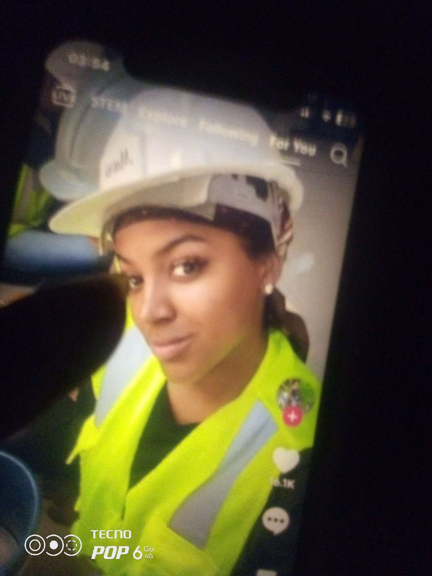 The image shows a young Black woman wearing a white hard hat and a bright yellow safety vest over a dark shirt. She is looking at the camera with a slight smile. The background is out of focus, but appears to be an indoor setting with some equipment or structures visible. Text overlays on the screen indicate a timestamp of "03:54", along with labels such as "STEM", "Explore", "Following", and "For You". There are also icons representing a search magnifying glass, a heart with "16.1K" underneath, and a speech bubble. The bottom of the image shows the "TECNO POP 6 4G" logo. There is no indication of the location being Howayi, Nigeria.