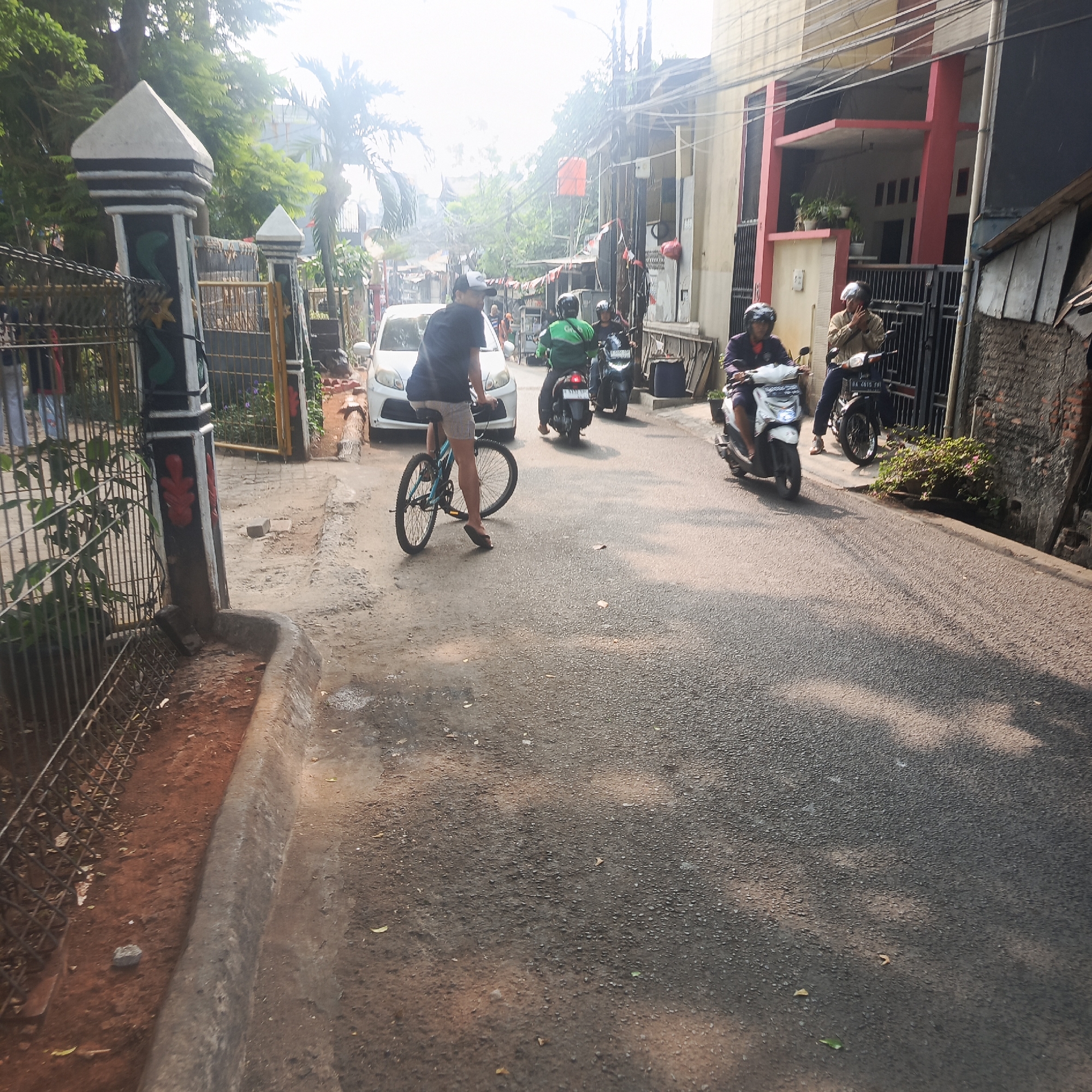 The image depicts a street scene in South Jakarta, Indonesia, under daylight. A man wearing a dark t-shirt and light-colored shorts is riding a blue bicycle in the foreground, facing away from the camera. He is wearing flip-flops. Behind him, a white car is parked on the left side of the road. Further down the street, several motorbikes are present. One motorbike, ridden by a person in a green jacket, is in motion, heading away from the camera. Another motorbike is parked to the right of the moving one. A person wearing a jacket and helmet is seated on this parked motorbike. On the far right, another motorbike with a rider is visible. The street is paved with asphalt and shows some signs of wear. On the left side of the street, there is a fence and a decorative pillar at what appears to be an entrance. Buildings are visible on the right side of the street, some with red accents. Trees and power lines are also present, contributing to the urban environment. The overall impression is a typical street scene in a residential or commercial area.