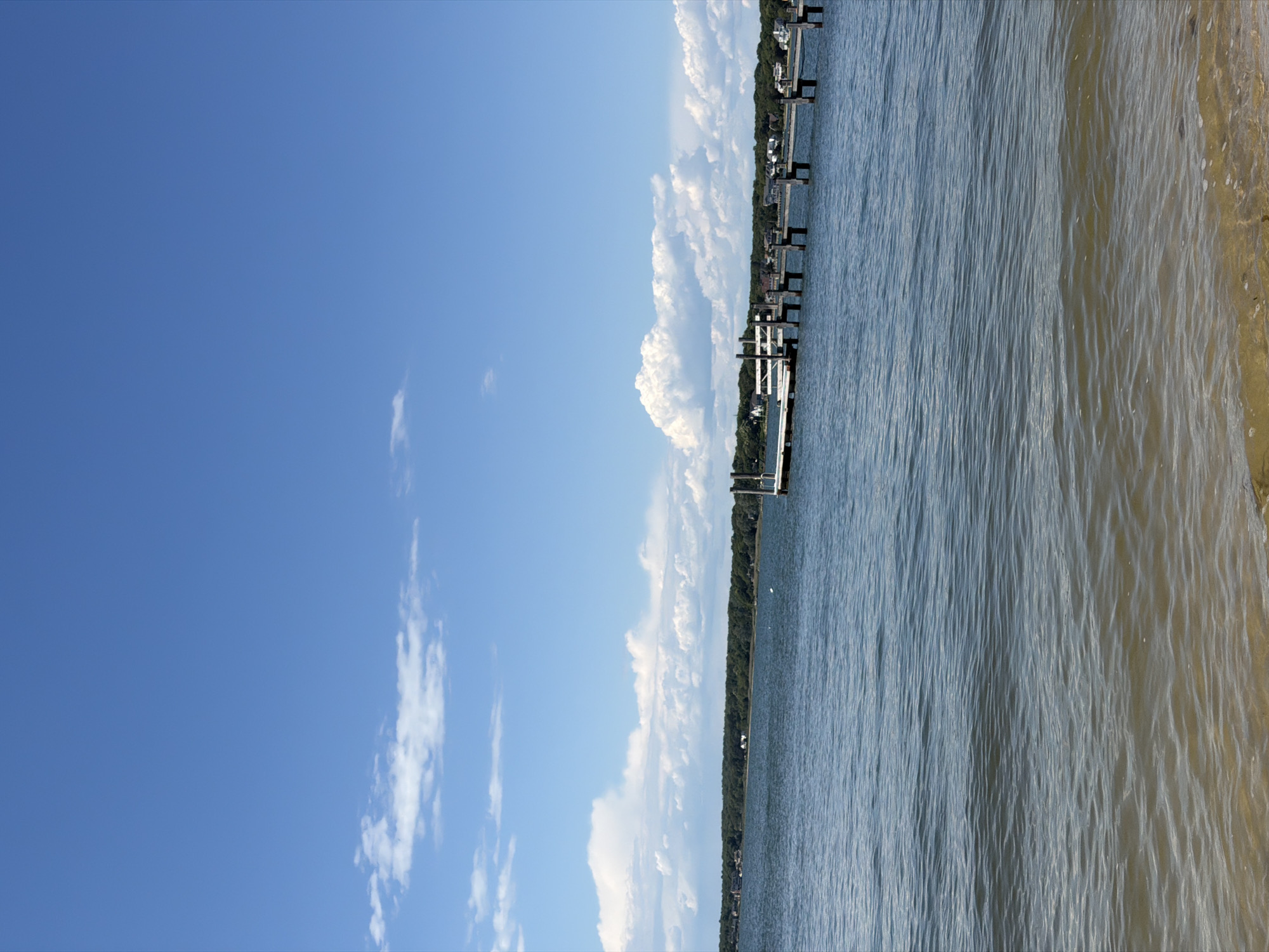 A vertical, wide shot captures a tranquil summer day at a waterfront in Hampton Bays, New York, USA. The scene is dominated by a vast expanse of clear blue sky dotted with fluffy white clouds. Below, a calm body of water reflects the sky, with gentle ripples disturbing the surface.
A wooden pier extends into the water from the right side of the frame, its weathered planks and sturdy pilings creating a linear element against the natural landscape. A small white boat is docked at the pier. The distant shoreline is densely lined with lush green trees, suggesting a wooded area, and interspersed with a few houses, indicating a residential neighborhood. The foreground shows the sandy edge of the water, with shallow, translucent waves lapping at the shore. The overall impression is one of peaceful, scenic waterfront living.