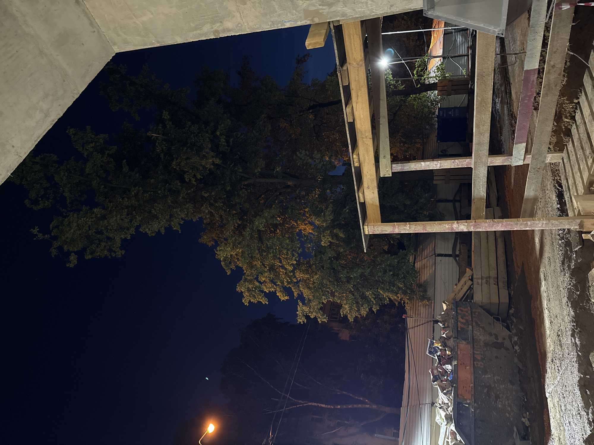 A nighttime outdoor construction scene is depicted, likely in Moscow, Russia, given the context provided. The image focuses on a large, orange dumpster overflowing with debris, positioned on a dirt surface. Adjacent to the dumpster are stacked concrete slabs, suggesting ongoing construction or demolition. In the background, construction fencing encloses the site, behind which trees with foliage are visible, some illuminated by artificial lighting. A streetlamp with an orange glow is also present, casting light on the scene and a nearby building facade. The foreground is dominated by wooden scaffolding and concrete structures, indicating part of the building under construction. The sky is dark, consistent with nighttime, and a sliver of the moon is visible. No people are present in the frame.