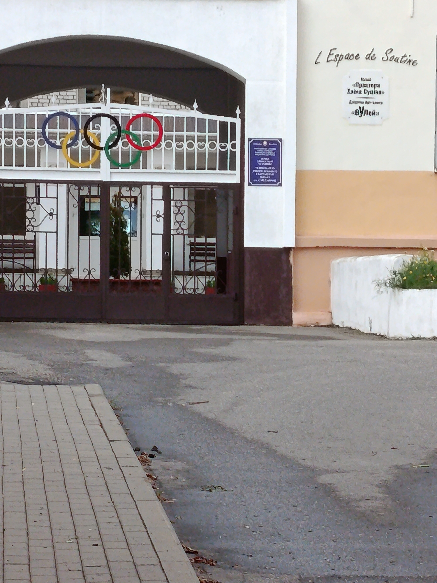 The image shows the exterior of a building in Смілавіцкі сельскі Савет, Belarus. The building has a white facade with a large arched entrance covered by a dark awning. In front of the entrance is a white metal gate decorated with ornamental scrollwork. Above the gate, the Olympic rings are displayed: a blue ring, a yellow ring, a black ring, a red ring, and a green ring, arranged from left to right.
To the right of the entrance, on a light beige wall, is a white sign with text in Cyrillic and Latin script. The Latin text reads "L'Espace de Soutine". Below this, the Cyrillic text appears to be a memorial plaque.
To the left of the entrance, a dark blue sign with white text is affixed to the white wall.
The gate itself is made of dark metal with intricate patterns. Through the gate, a courtyard is visible with two dark benches on either side and a small evergreen tree in a planter in the center.
The foreground shows a grey asphalt surface and a grey brick sidewalk on the left side of the image. A white concrete planter with some green vegetation is visible on the far right side of the building. The overall scene suggests an entrance to a public or cultural institution.