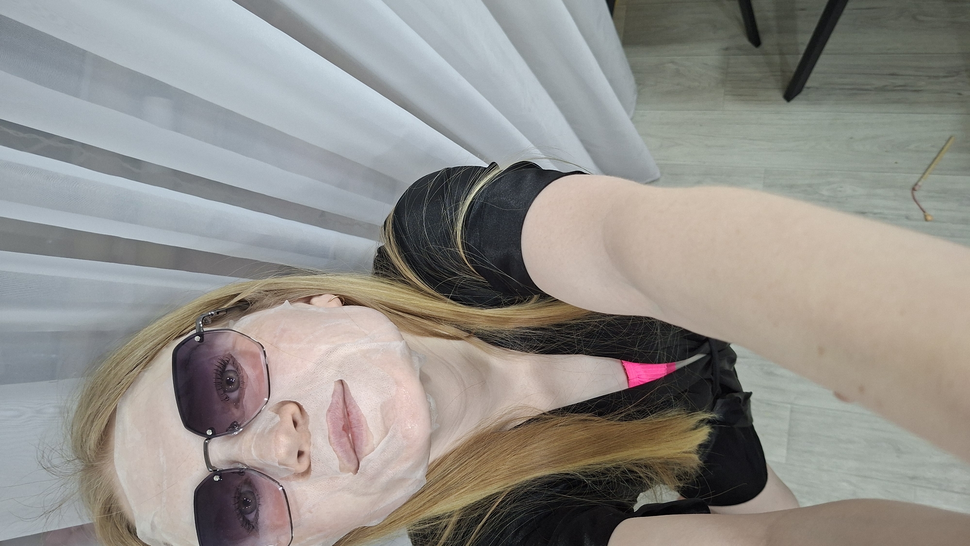A woman is pictured indoors, possibly in a private residence or spa-like setting. She lies on her back, wearing a facial mask and sunglasses. Her hair is blonde. She wears a black top with a glimpse of pink underneath. The background includes a white curtain or blind and a partial view of a black chair leg. The floor is light-colored and appears to be made of planks. The location is in the city of Chaikovsky, Russia. The photograph is likely a selfie or taken from a low angle.