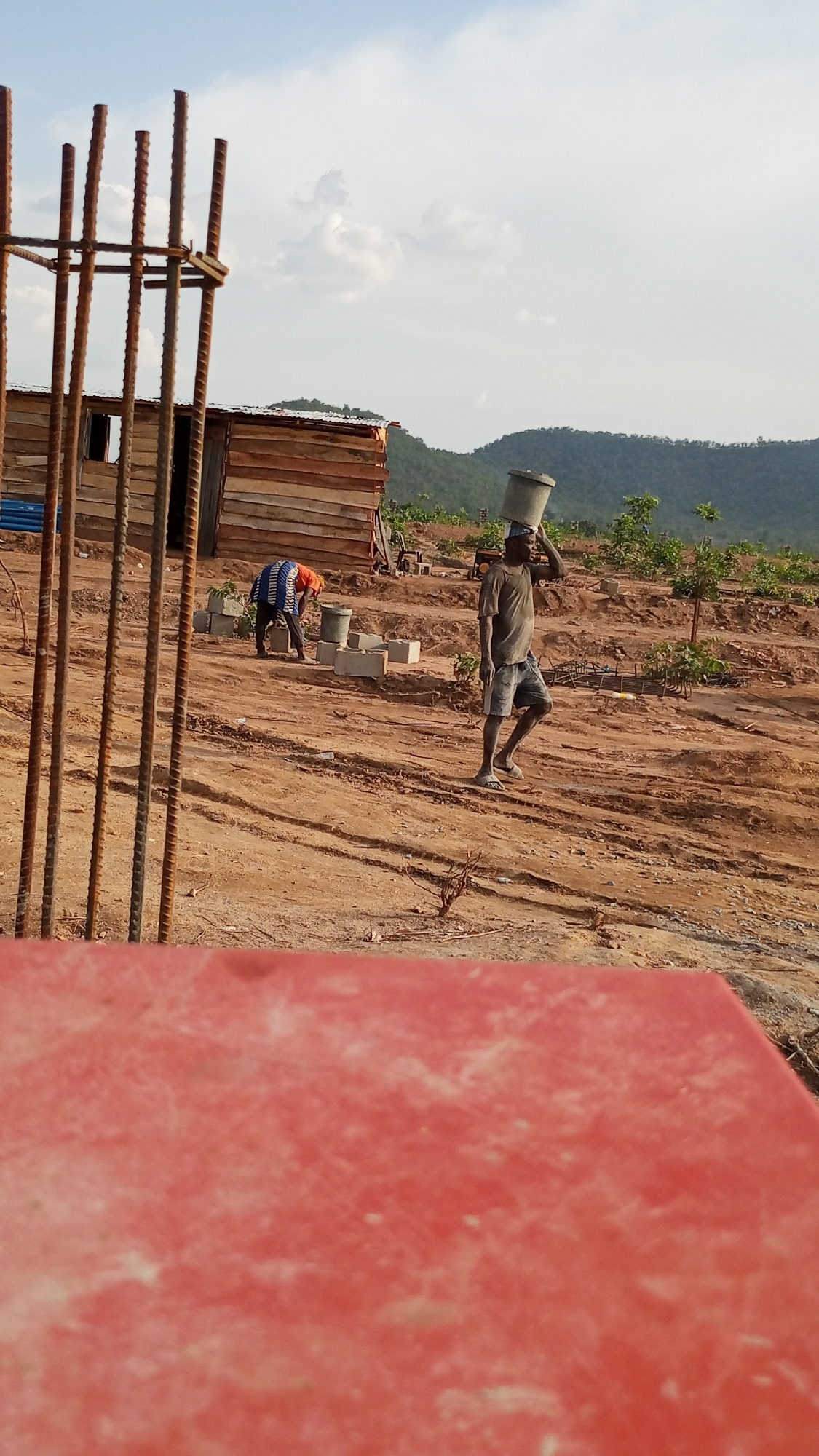 In Zango-ndaji, Nigeria, the scene shows a construction site with a wooden structure in the background. Two men are present; one carries a bucket on his head, while the other is working near a pile of cinder blocks. Rebar stands vertically in the foreground, and a red object obstructs the lower portion of the image. The ground is dirt, and there are hills in the distance.