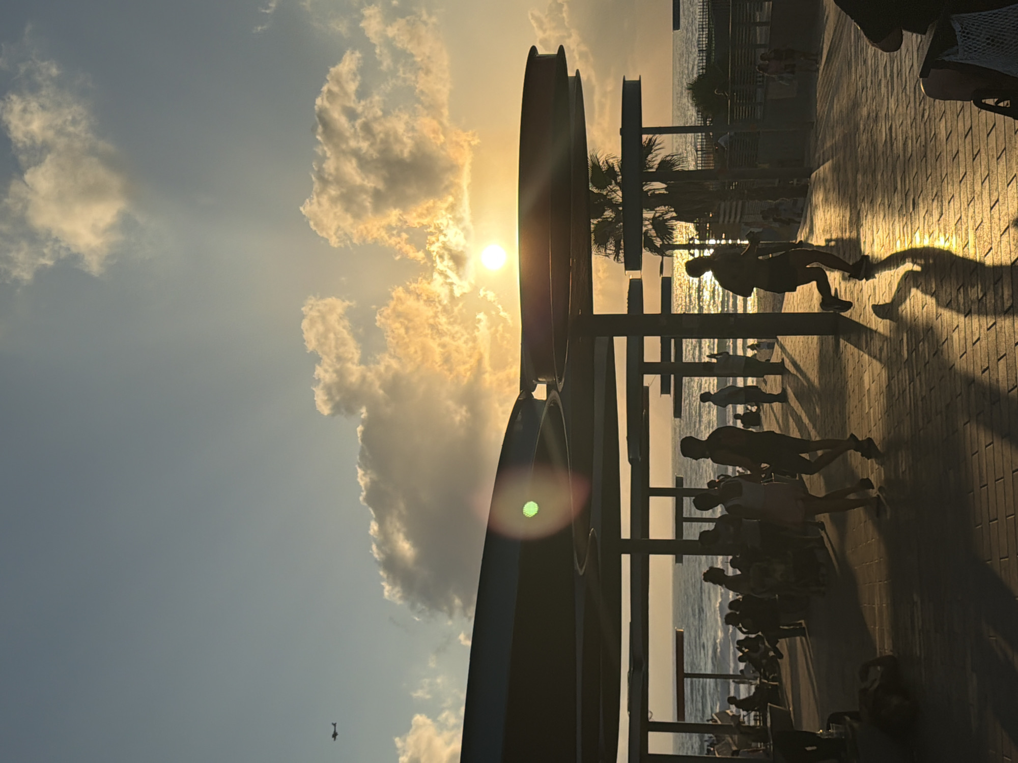 The image depicts a sunset scene at the Bat Yam promenade in Israel. The sun is low on the horizon, casting a warm glow across the sky and the water. Silhouetted figures are scattered throughout the scene. In the foreground, several people are walking and standing under a modern, curved canopy structure. To the left, a small airplane is visible in the sky. Further down the promenade, more people are seen sitting and walking. Palm trees add a tropical touch to the environment. The ocean is calm, with gentle waves reflecting the golden light of the setting sun. The promenade is paved with interlocking bricks, creating a patterned walkway. The overall atmosphere is one of leisure and relaxation, with people enjoying the evening by the sea.