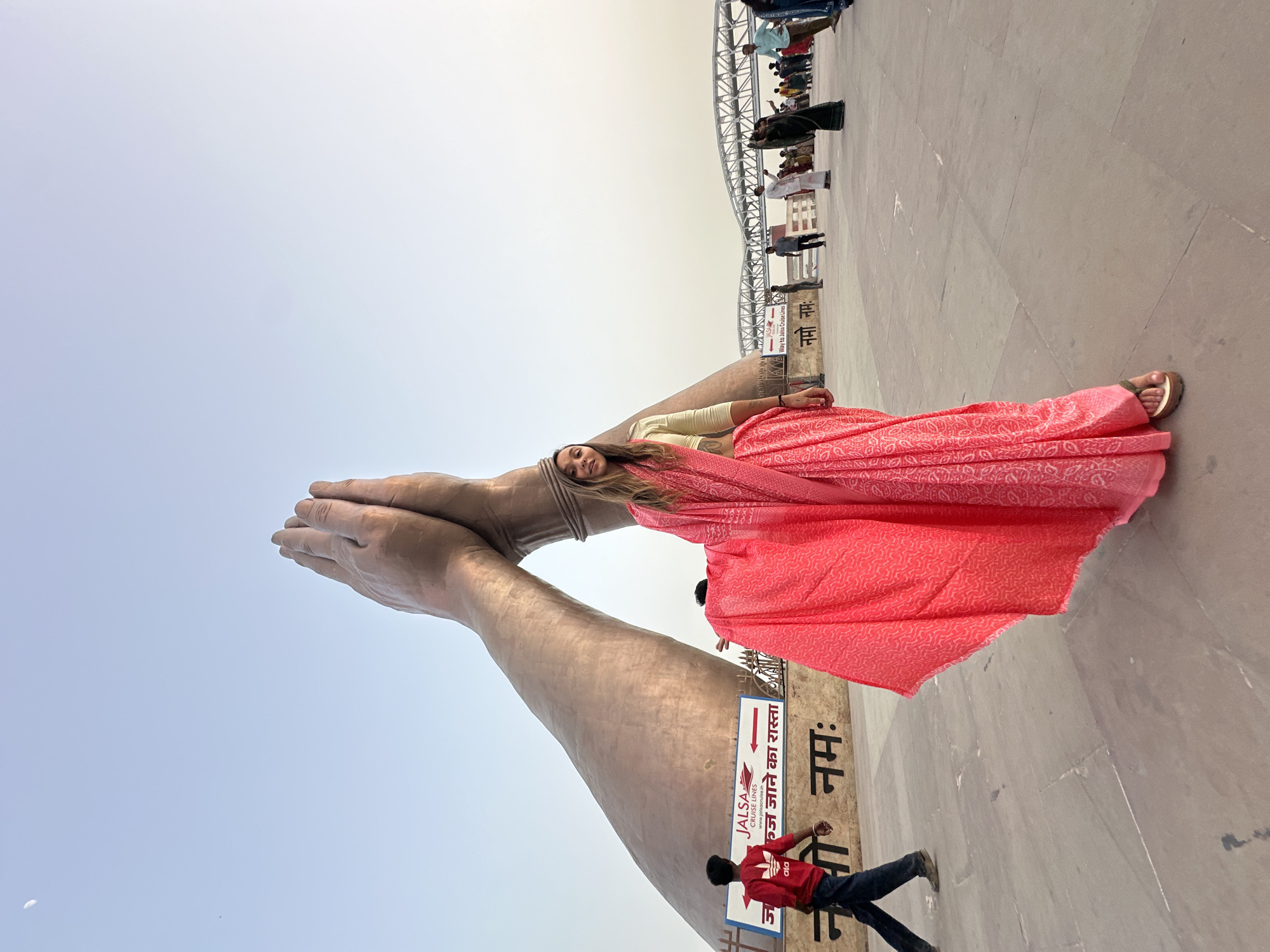 A medium shot captures a woman in a coral-colored sari positioned between a pair of massive, sculpted hands and a pedestrian walkway near the Jalsa Cruise Lines in Mughalsarai, India. The woman, with shoulder-length blonde hair, is angled slightly towards the camera. She wears a light-colored blouse beneath her sari and sandals on her feet. The large, metallic hands are positioned vertically in a gesture of prayer or greeting. Behind her is a flat, paved area with a sign pointing the "Way to Jalsa Cruise Lines" in both English and Hindi. A man in a red shirt and jeans is walking near the sign. In the background, a line of people stretches along a bridge-like structure. The sky is overcast, providing a muted backdrop.