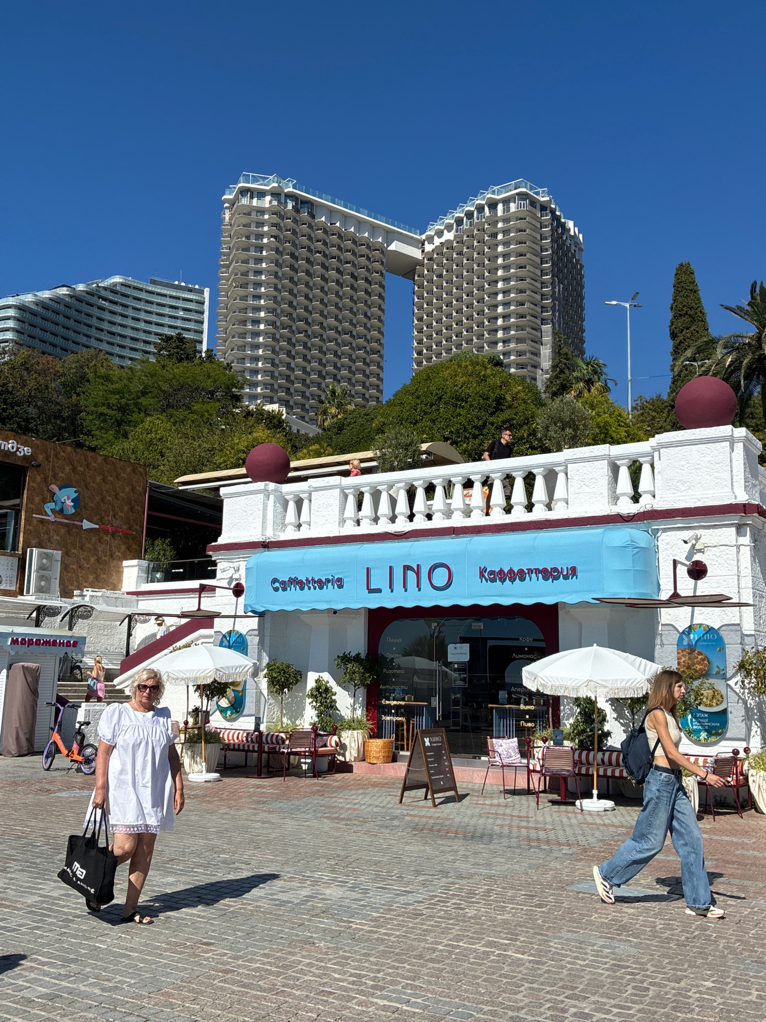 The image depicts a bright, sunny day in Sochi, Russia, likely near the coastline, given the presence of palm trees and the open, outdoor setting. In the background, two imposing, modern high-rise apartment buildings with balconies dominate the skyline, one of which appears to be connected to the other by a skybridge. In the foreground, a white, two-story building with red trim serves as a cafe or restaurant named "LINO" with "Кафеттерия" (Caffetteria) written in Russian above the entrance. White umbrellas provide shade to outdoor seating areas.
Several people are visible. A woman in a white dress and sunglasses walks from left to right, carrying a black tote bag. To her right, a young woman in a white tank top and baggy jeans walks in the same direction, wearing a backpack. Further in the background, a man in a dark shirt and sunglasses stands on the balcony of the white building, looking out. Another person, a child, is visible on the same balcony. A purple and orange scooter is parked to the left of the main building, next to a sign with Russian text, possibly indicating "Morozhenoe" (Ice Cream). The ground is paved with cobblestones. Lush green trees and vegetation surround the buildings, suggesting a well-maintained urban environment. The overall impression is one of a vibrant resort town on a clear day.