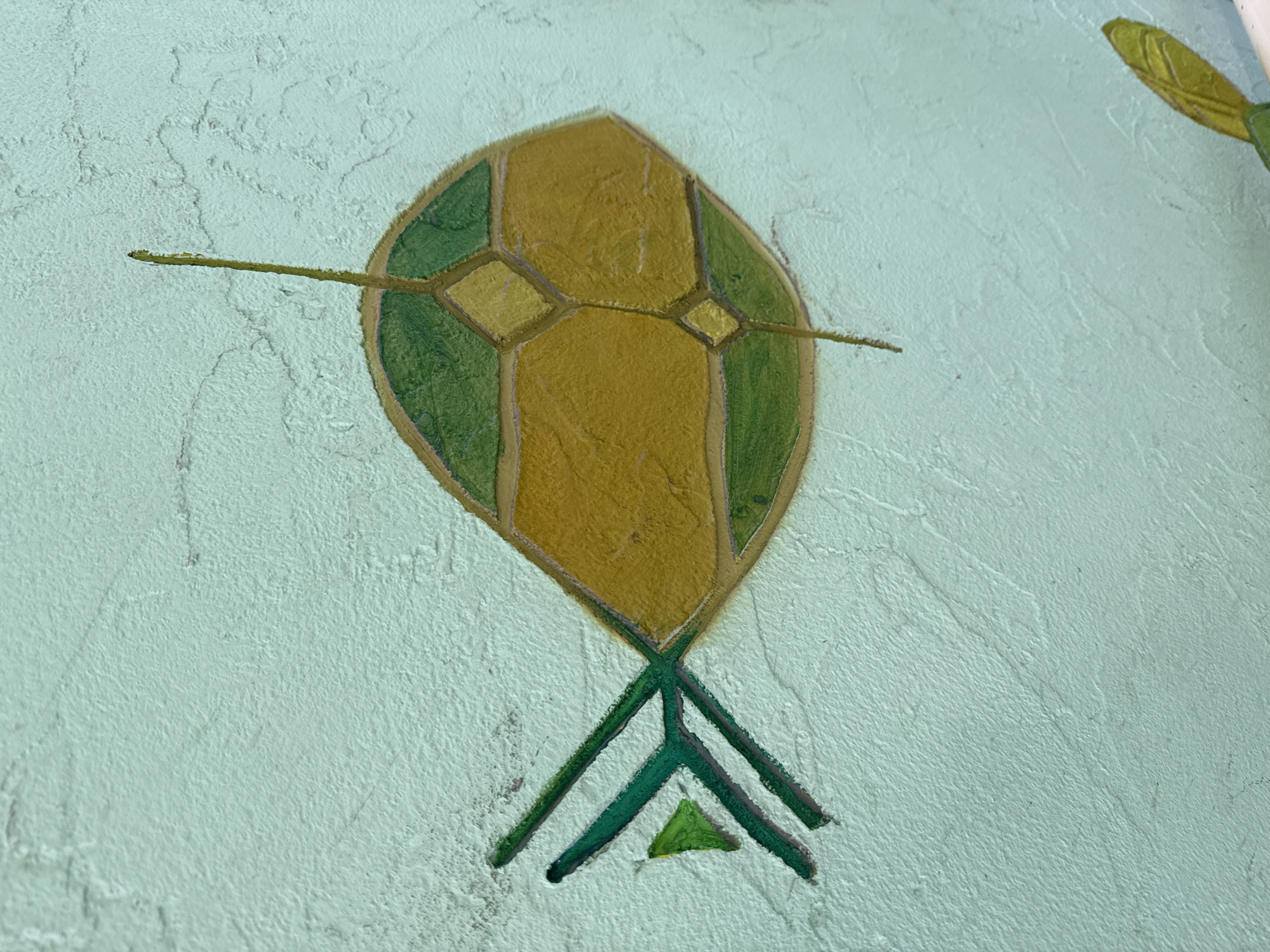 The image shows a stylized, painted representation of a kite on a textured, light blue surface, possibly a wall. The kite is predominantly yellow with green segments on either side of the upper portion. A horizontal green line passes through the kite's center. At the bottom, three green lines converge, ending above a small green triangle. The location is unknown.