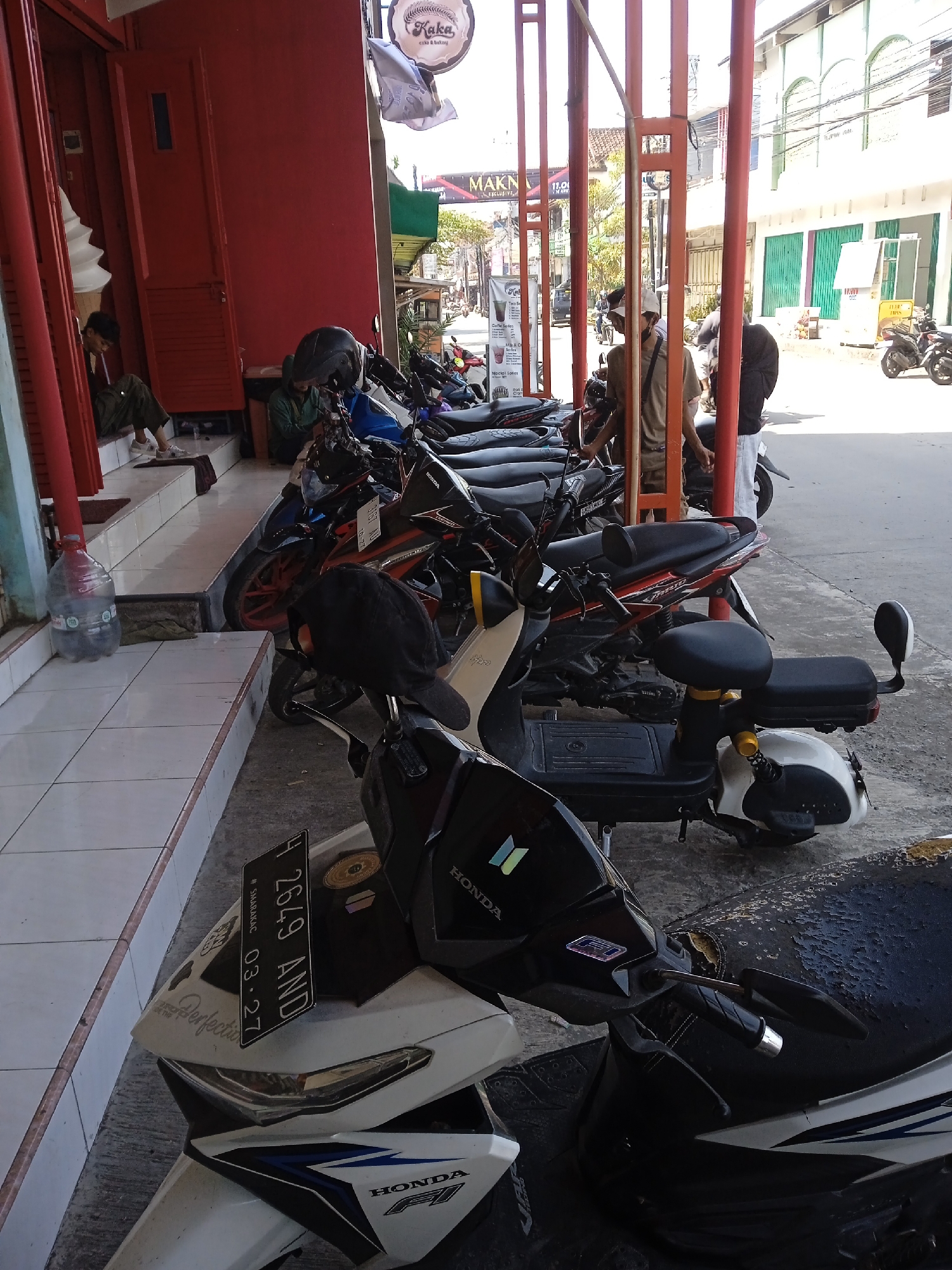 This image depicts a street scene in Pandean, Indonesia, characterized by a row of parked motorbikes along a building's exterior. The motorbikes are varied, with one particularly visible in the foreground showcasing a license plate reading "F 2649 AND #SMARAKAC 03.27". A Honda emblem is discernible on the front of the motorcycle.
The environment is urban, with red-painted building exteriors and a nearby establishment named "Kaka Cake & Bakery," identifiable by a sign. Further down the street, "MAKN EXCLUSIVE" is visible. Several individuals are present, including people standing near the motorbikes and one seated on the building's steps. A large plastic water bottle is placed near the steps. The scene is filled with natural sunlight, indicating daytime. An electric scooter with a peculiar seating arrangement is visible among the motorbikes.